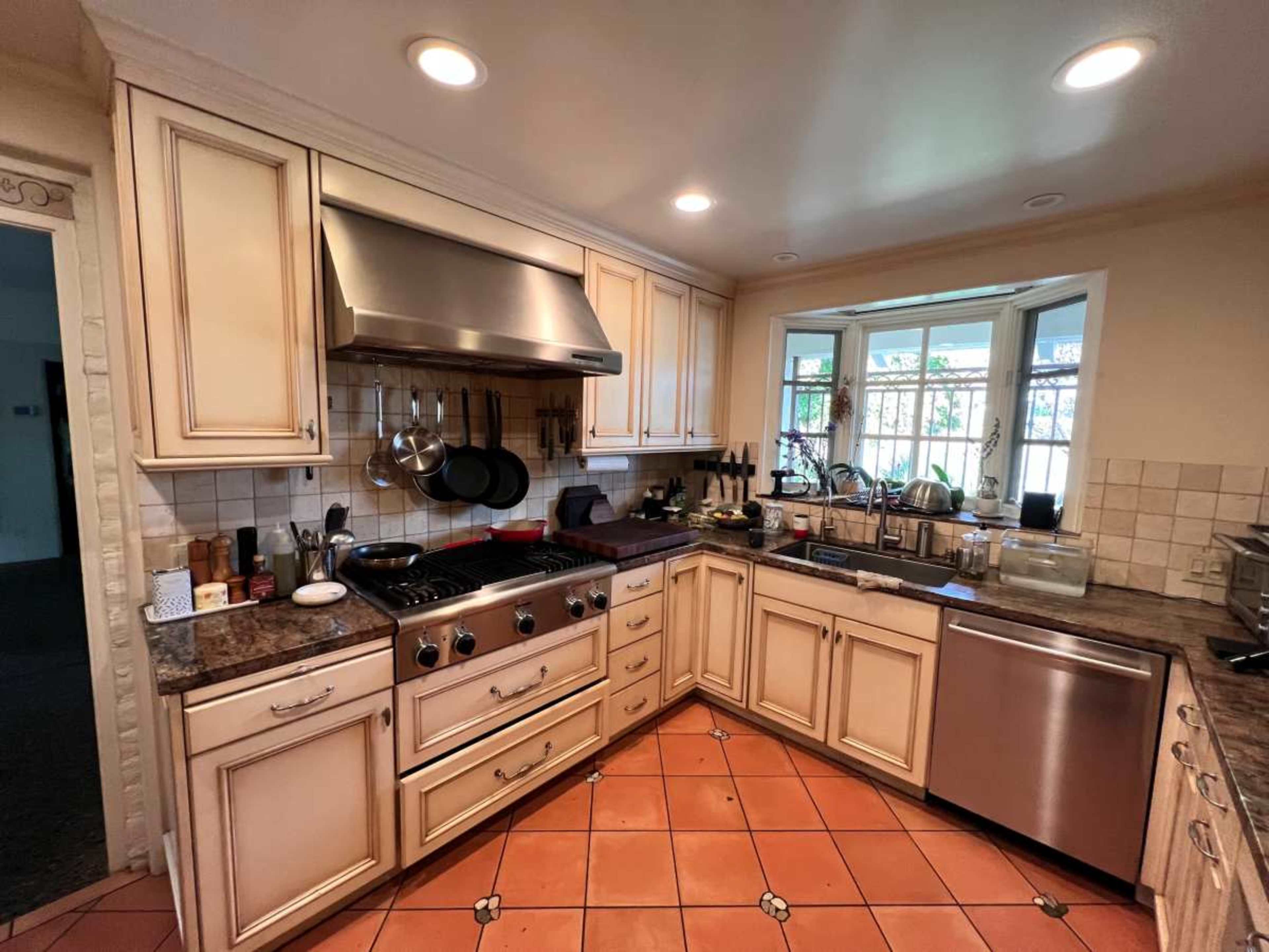 The image shows a kitchen with cream-colored cabinets, a stainless steel oven hood, and a tiled floor, featuring various kitchen utensils and appliances on the countertops.