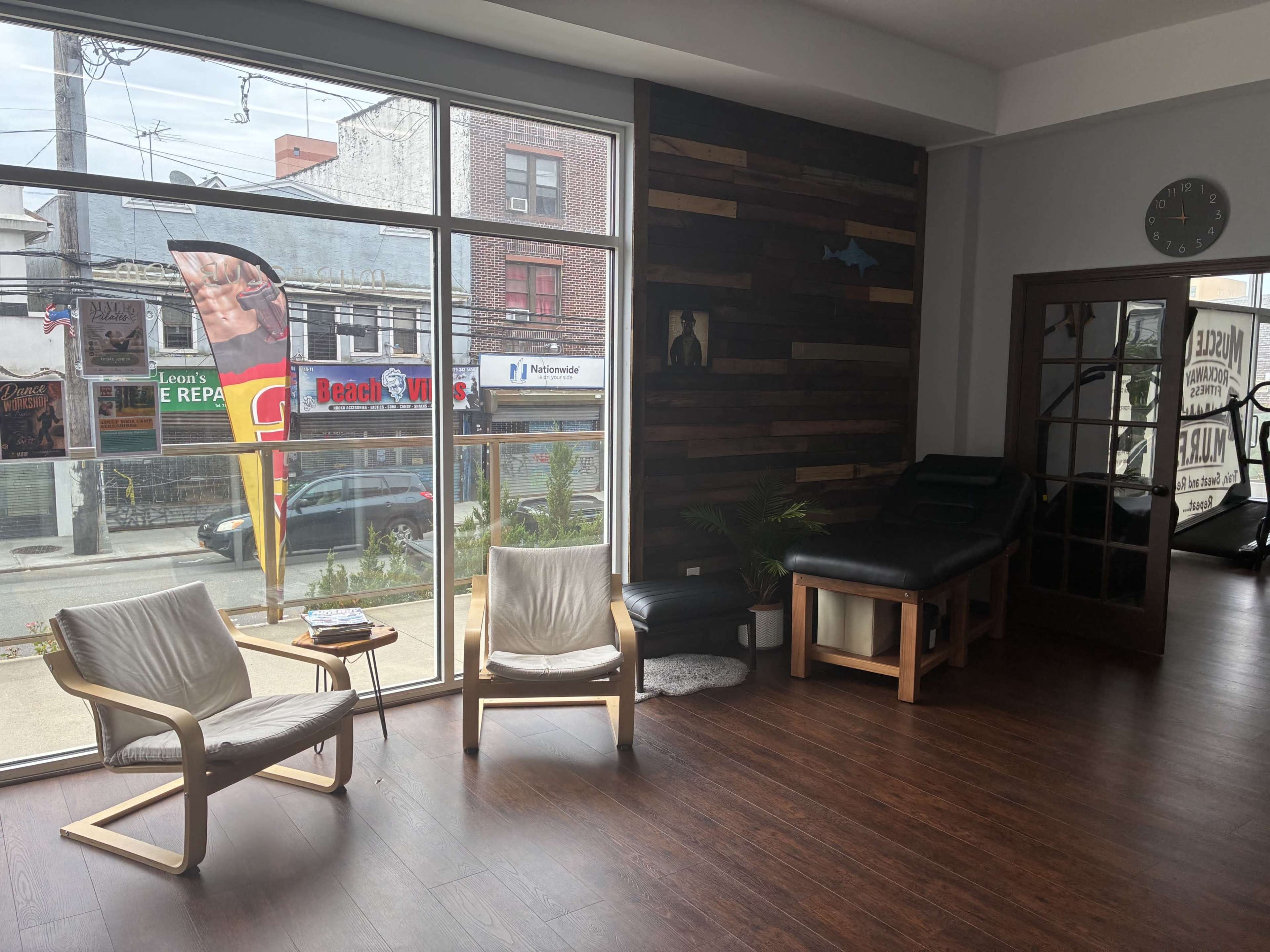 The image shows a spacious interior of a business with wooden accents, featuring two chairs, a side table, and a faintly visible treatment area through a doorway.