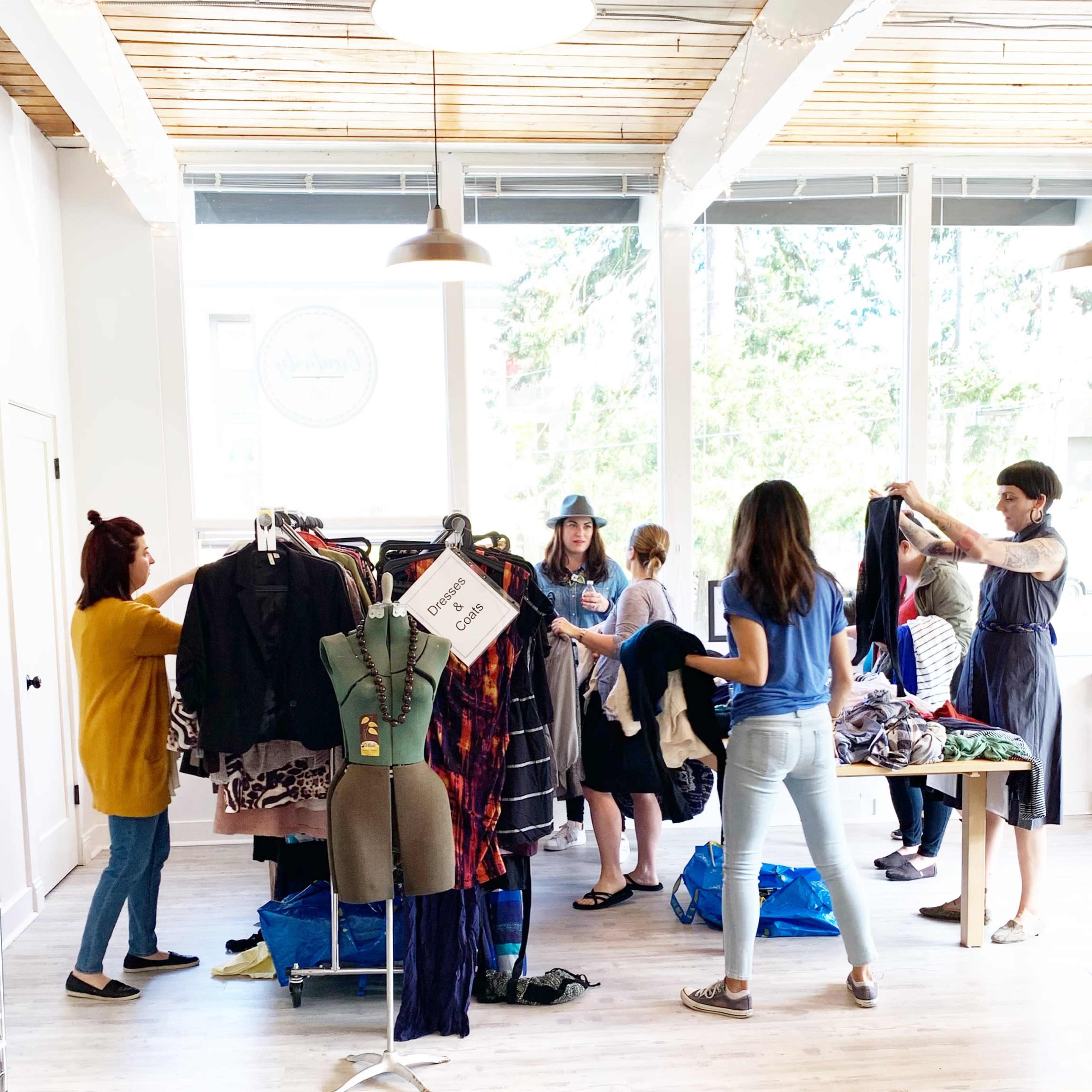 A group of people is sorting through clothes on racks and tables in a bright room with large windows.