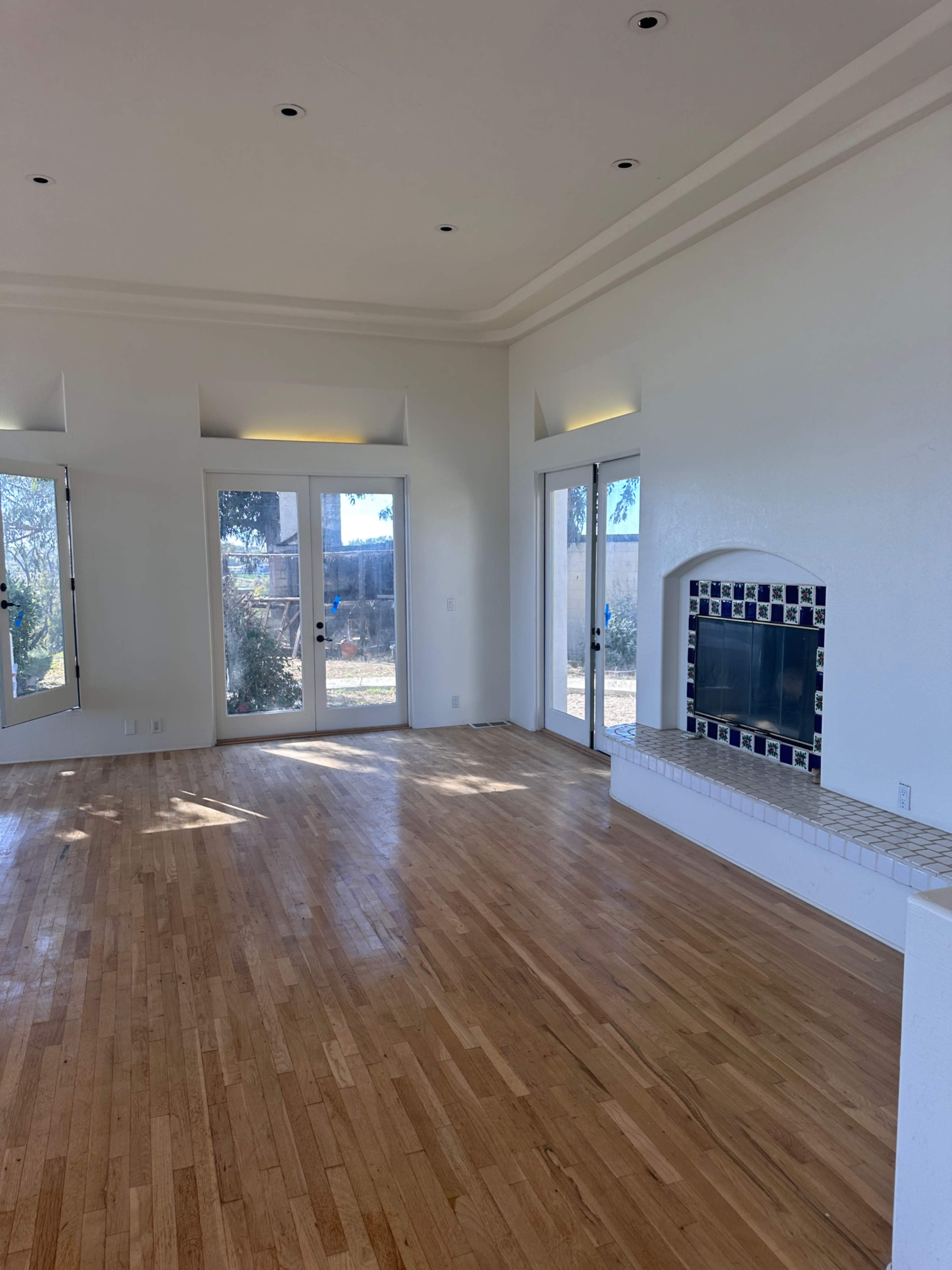 The image depicts a spacious living room with wooden flooring, large windows, and a white fireplace.