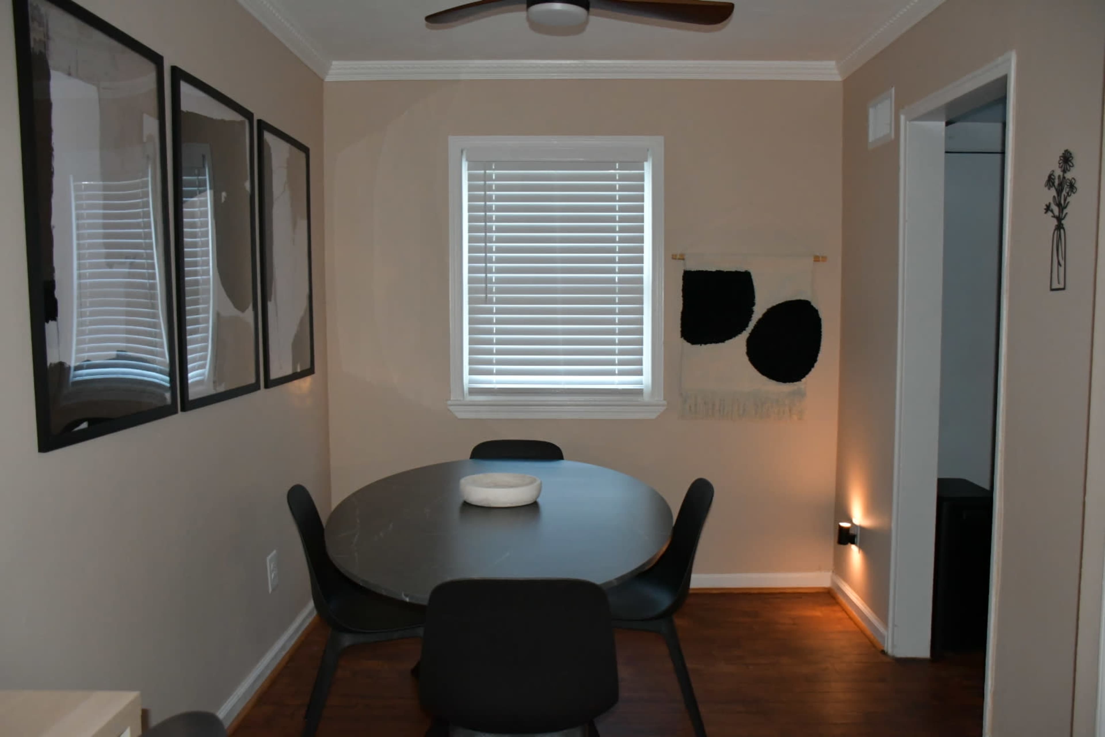The image shows a dining area featuring a round black table surrounded by four black chairs, with framed artwork on the walls and a window with white blinds.