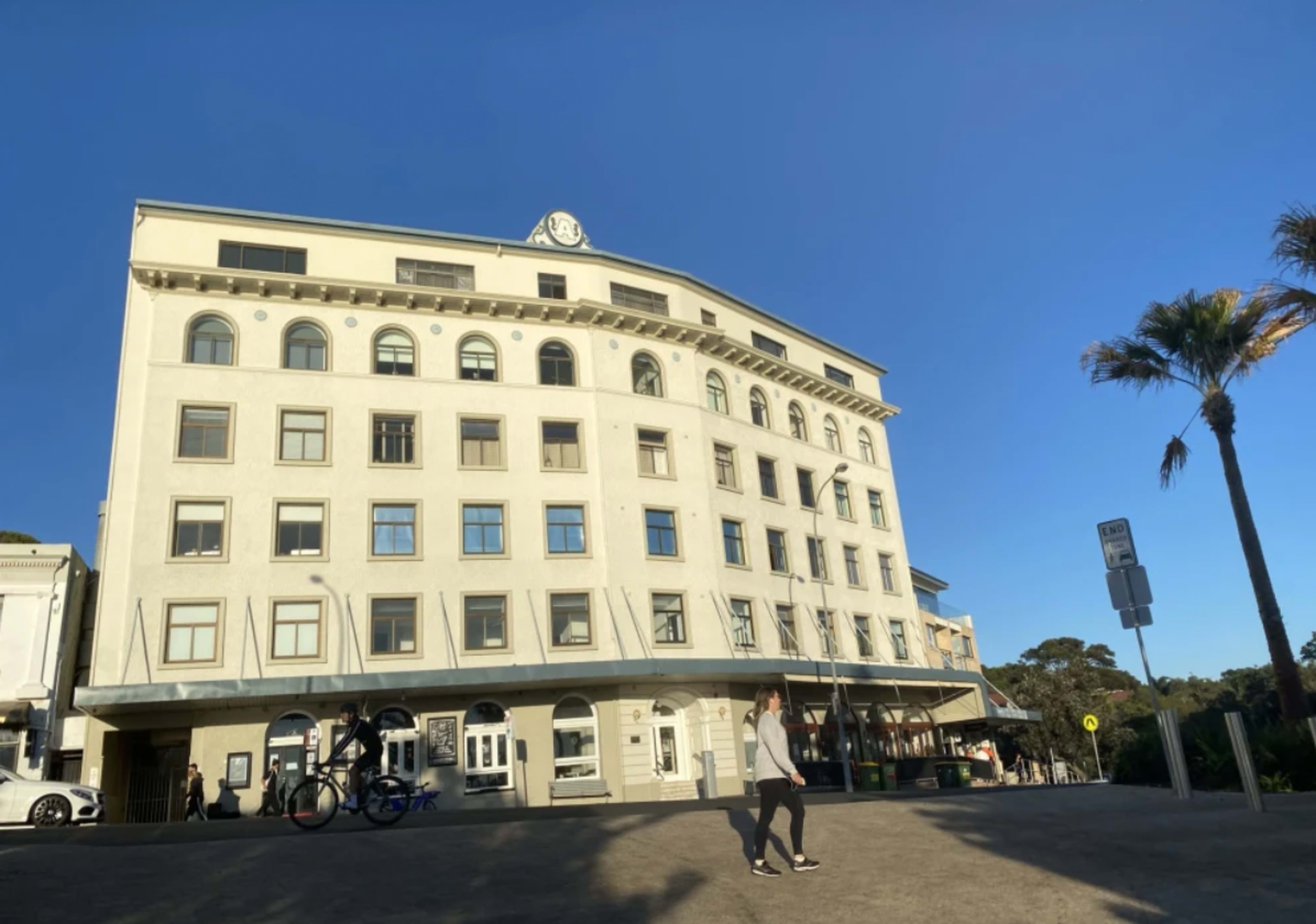A person walks past a historic building with a clock on the roof, located in a coastal area with palm trees.