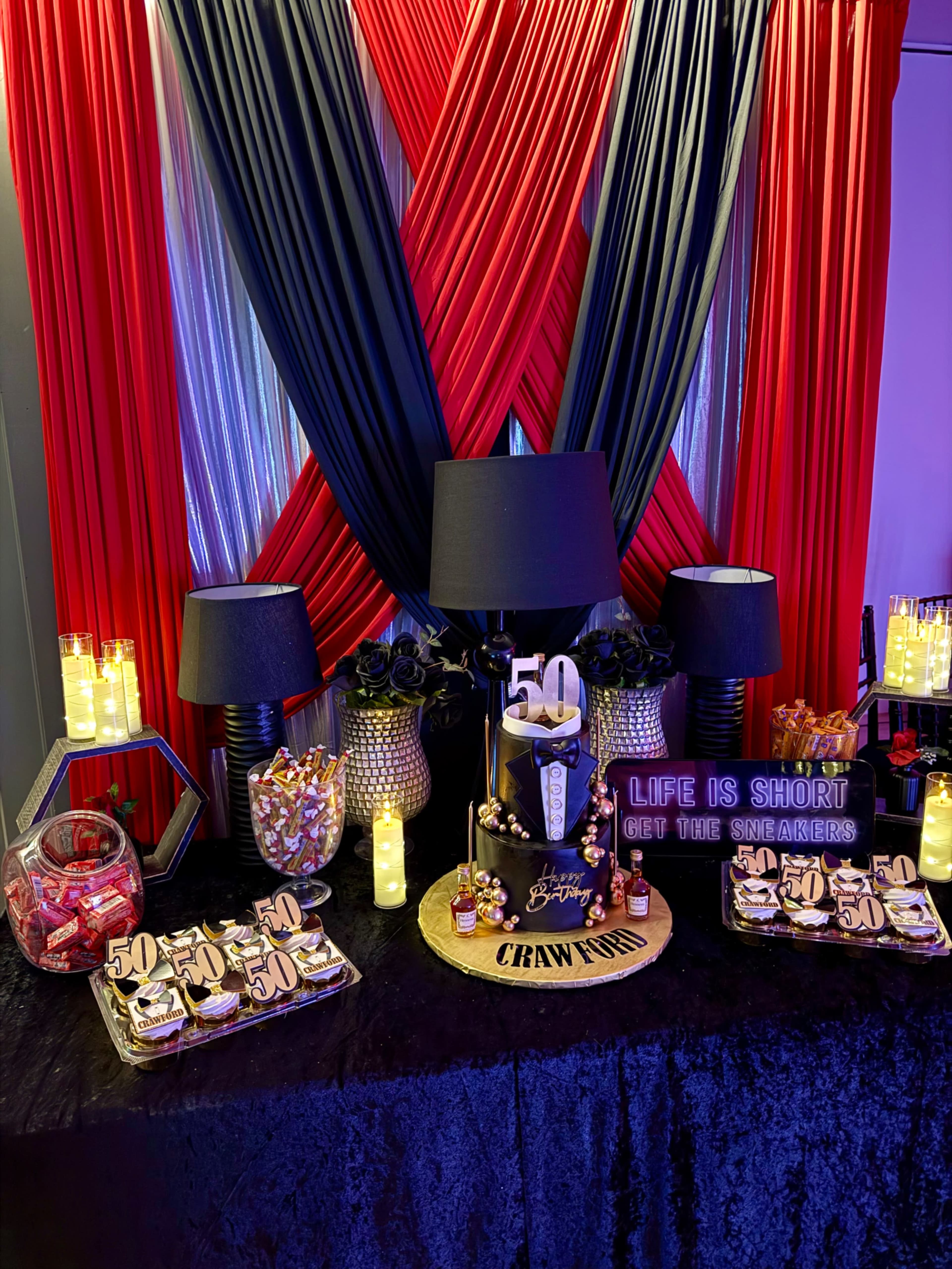 A decorated table features a 50th birthday theme with a black and red backdrop, snacks, candy, and a sign that reads, "LIFE IS SHORT GET THE SNEAKERS."