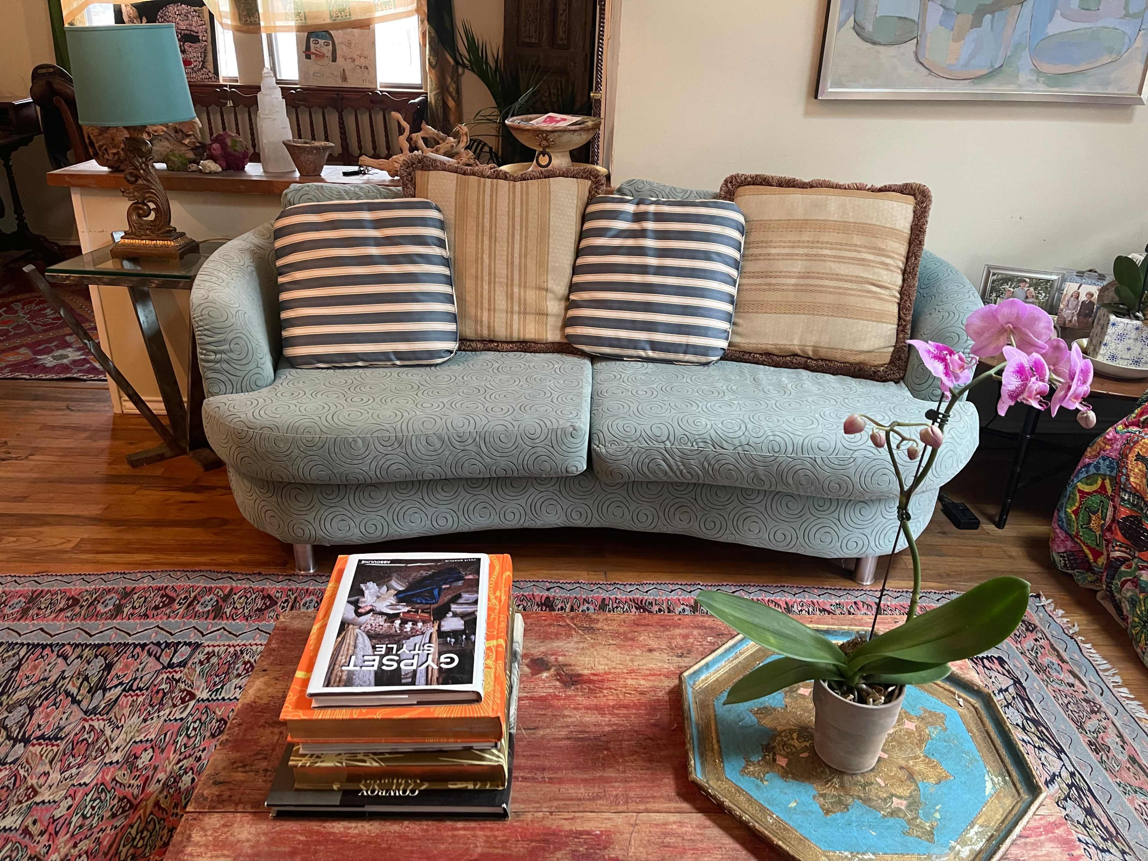 A light blue sofa with striped and textured decorative pillows, positioned in a cozy living space with a patterned rug and a small table displaying books and a potted orchid.