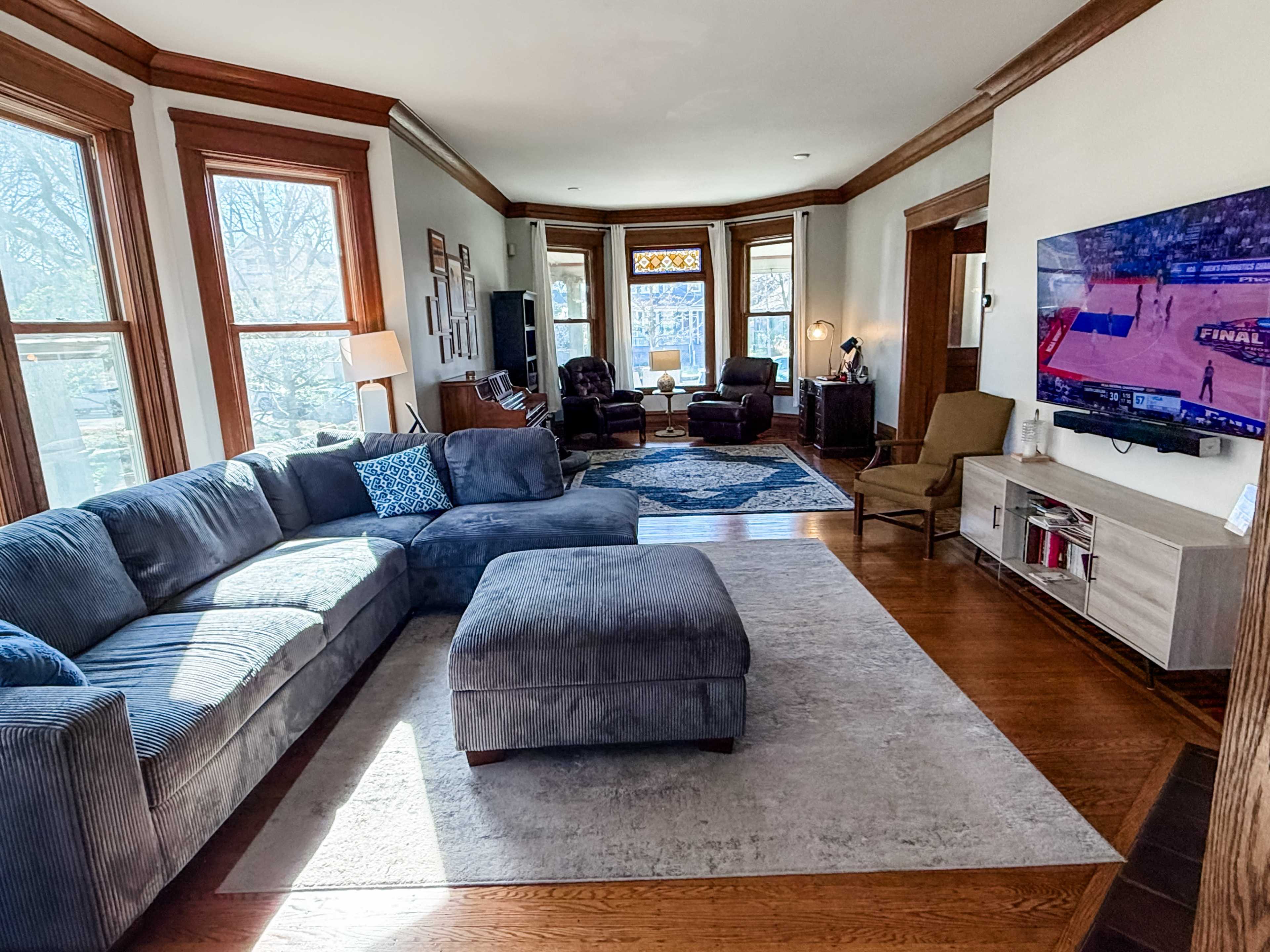 The image shows a bright living room with large windows, a sectional sofa, an ottoman, and a television displaying a basketball game.