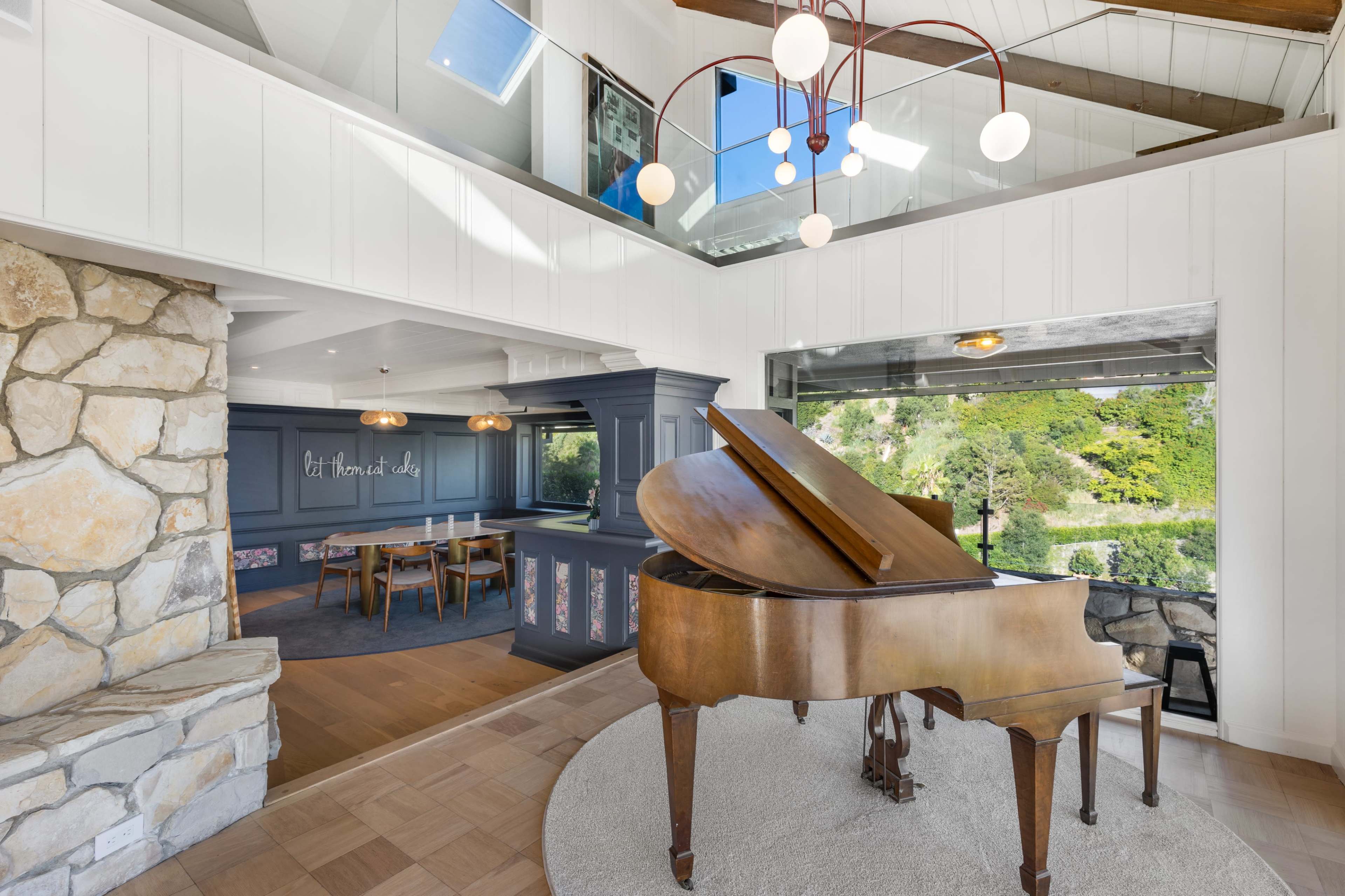 The image shows a spacious interior with a grand piano in the foreground, adjacent to a dining area and a large window that opens to an outdoor view.