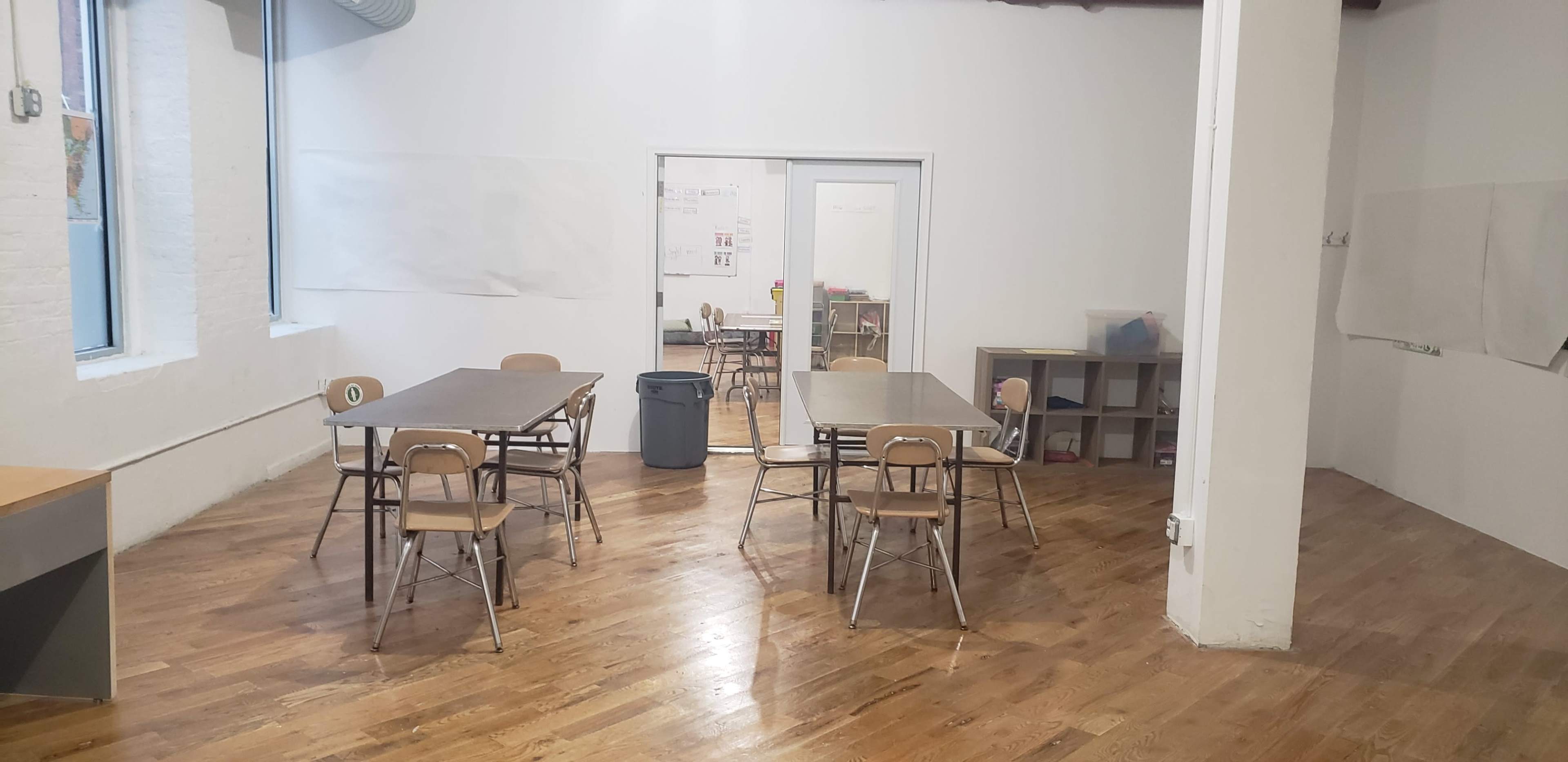 The image shows an empty classroom with several wooden tables and chairs arranged around a hardwood floor, along with a storage shelf and a trash can.