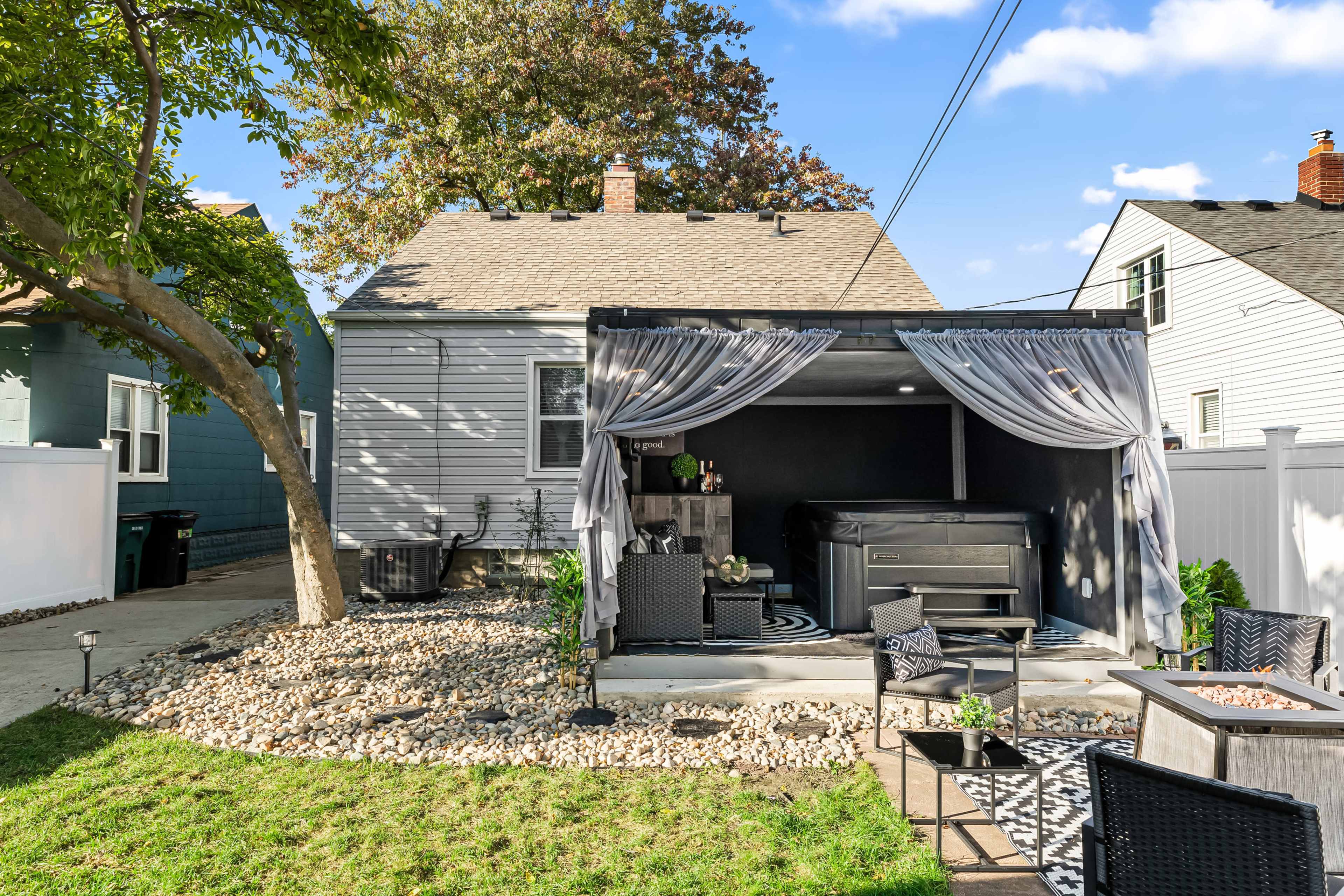The image depicts a backyard with a covered patio featuring a hot tub, surrounded by decorative stones and plants, along with outdoor seating arrangements.