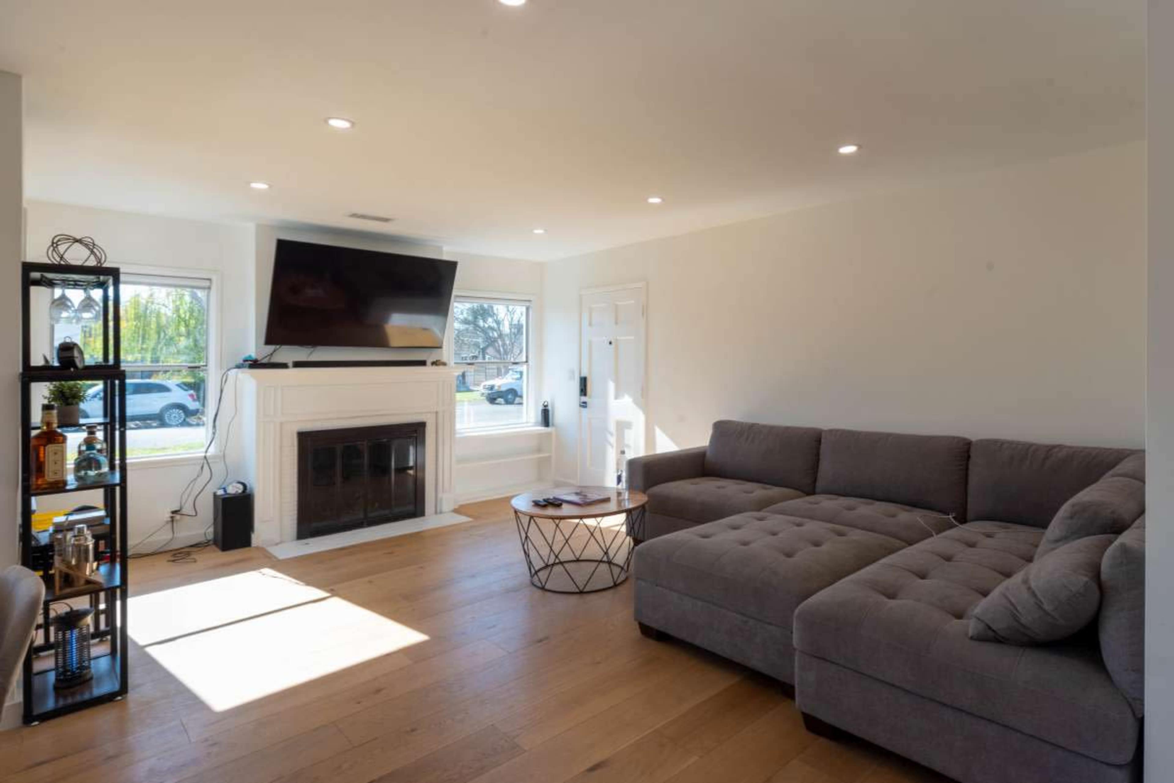 A modern living room features a sectional sofa, a coffee table, a wall-mounted TV, and a fireplace, with large windows allowing natural light to enter.