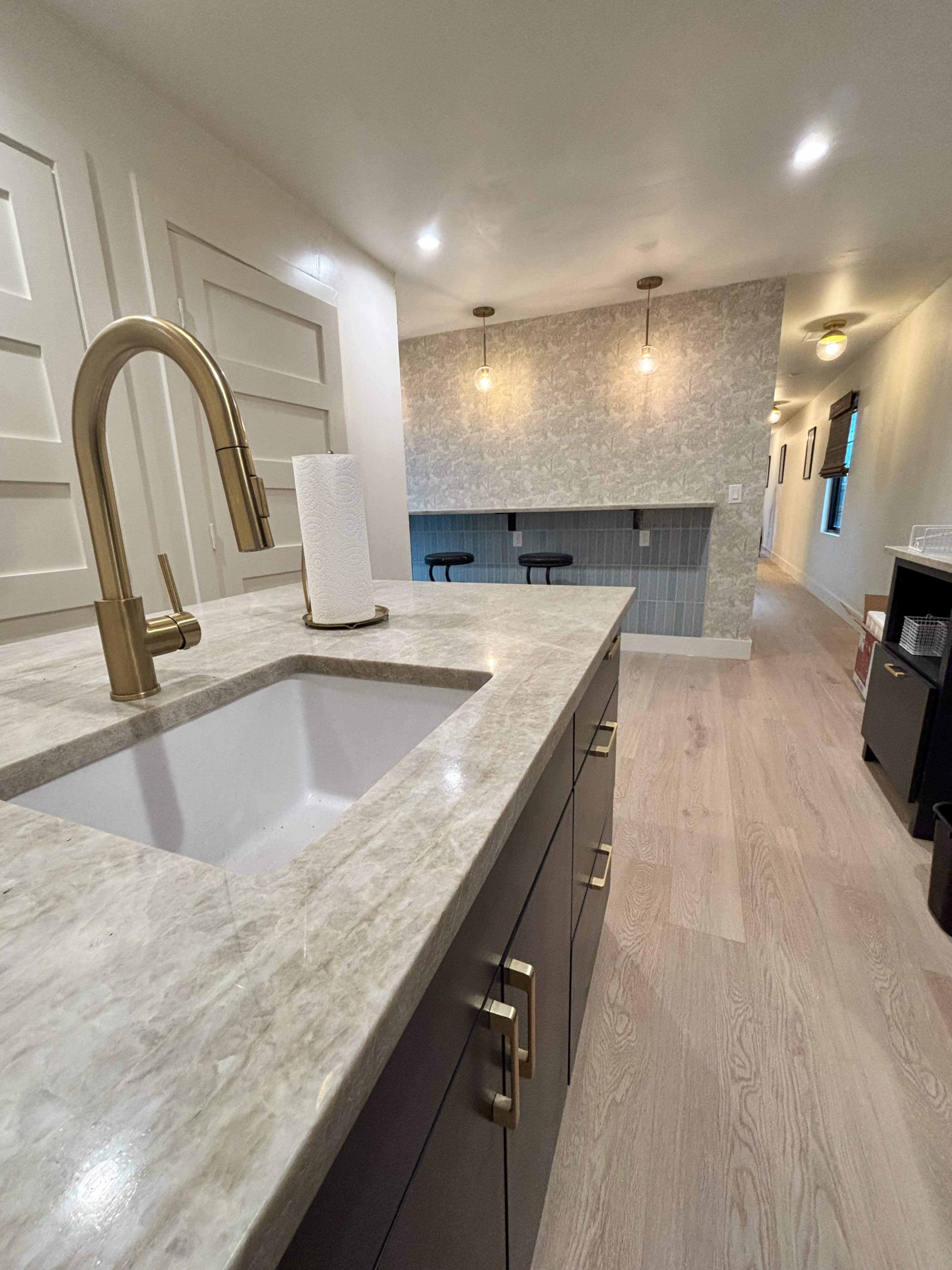 The image shows a well-lit interior space with a kitchen island featuring a sink and a gold faucet, leading to a distant seating area with pendant lights and a patterned wall.