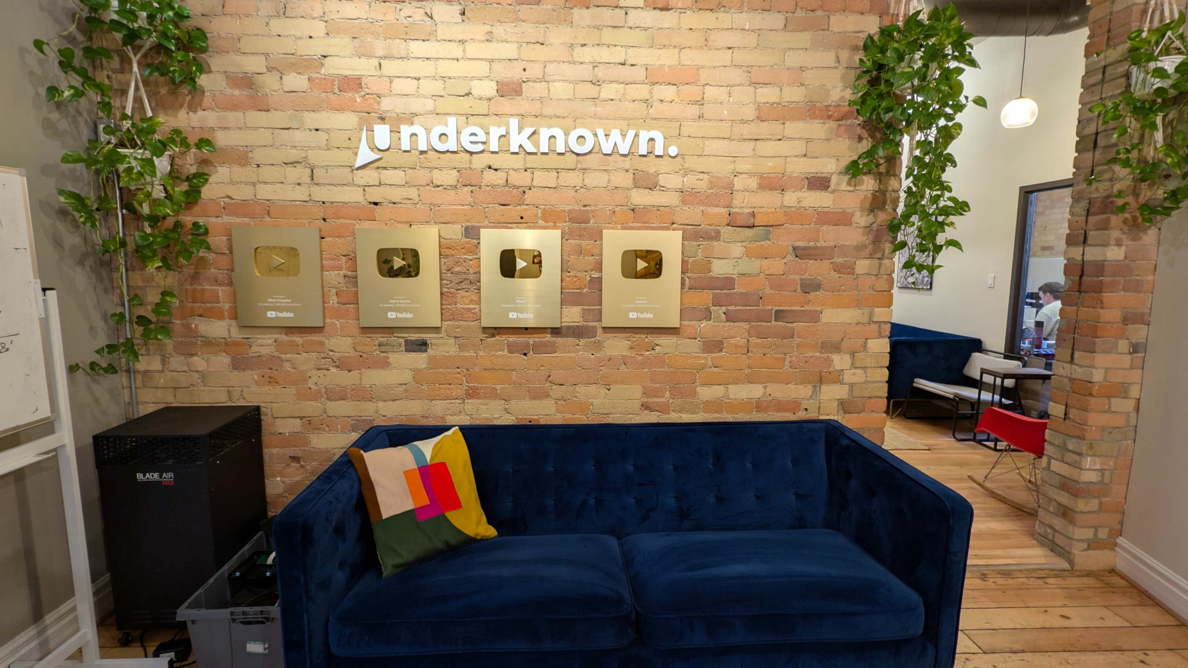 The image shows a cozy lounge area with a blue couch against a brick wall, decorated with four gold YouTube play buttons and green plants.