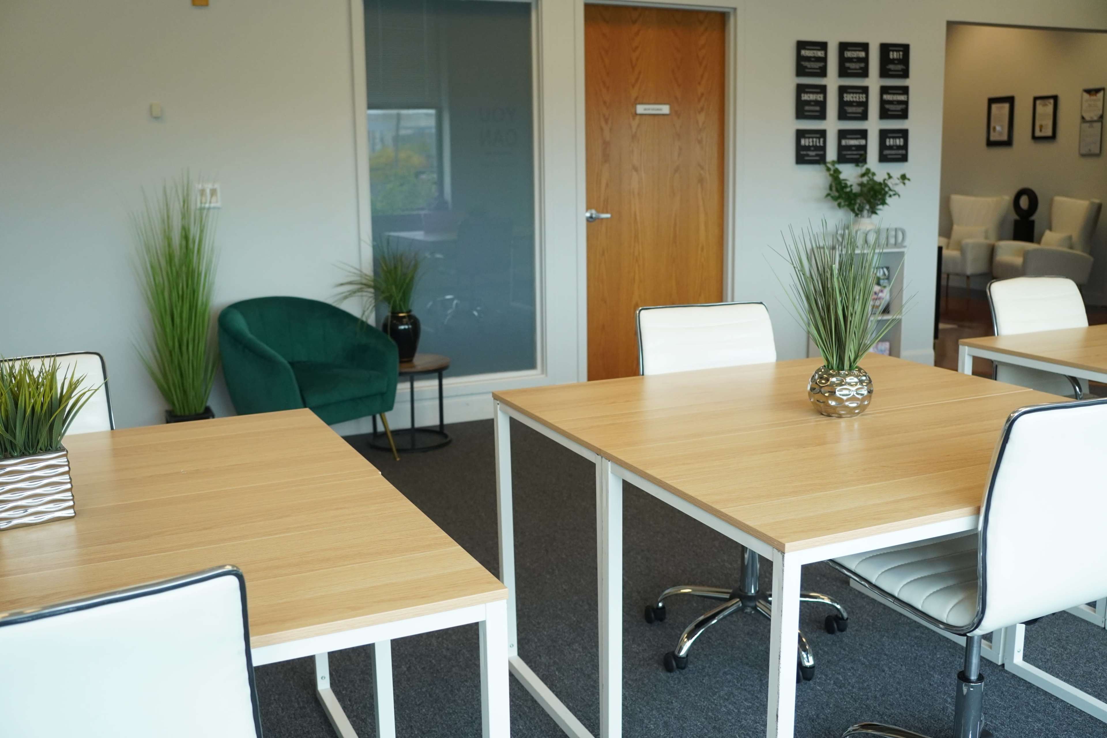 A modern office space features two wooden tables with white chairs, a green armchair in the corner, and plants positioned on the tables.