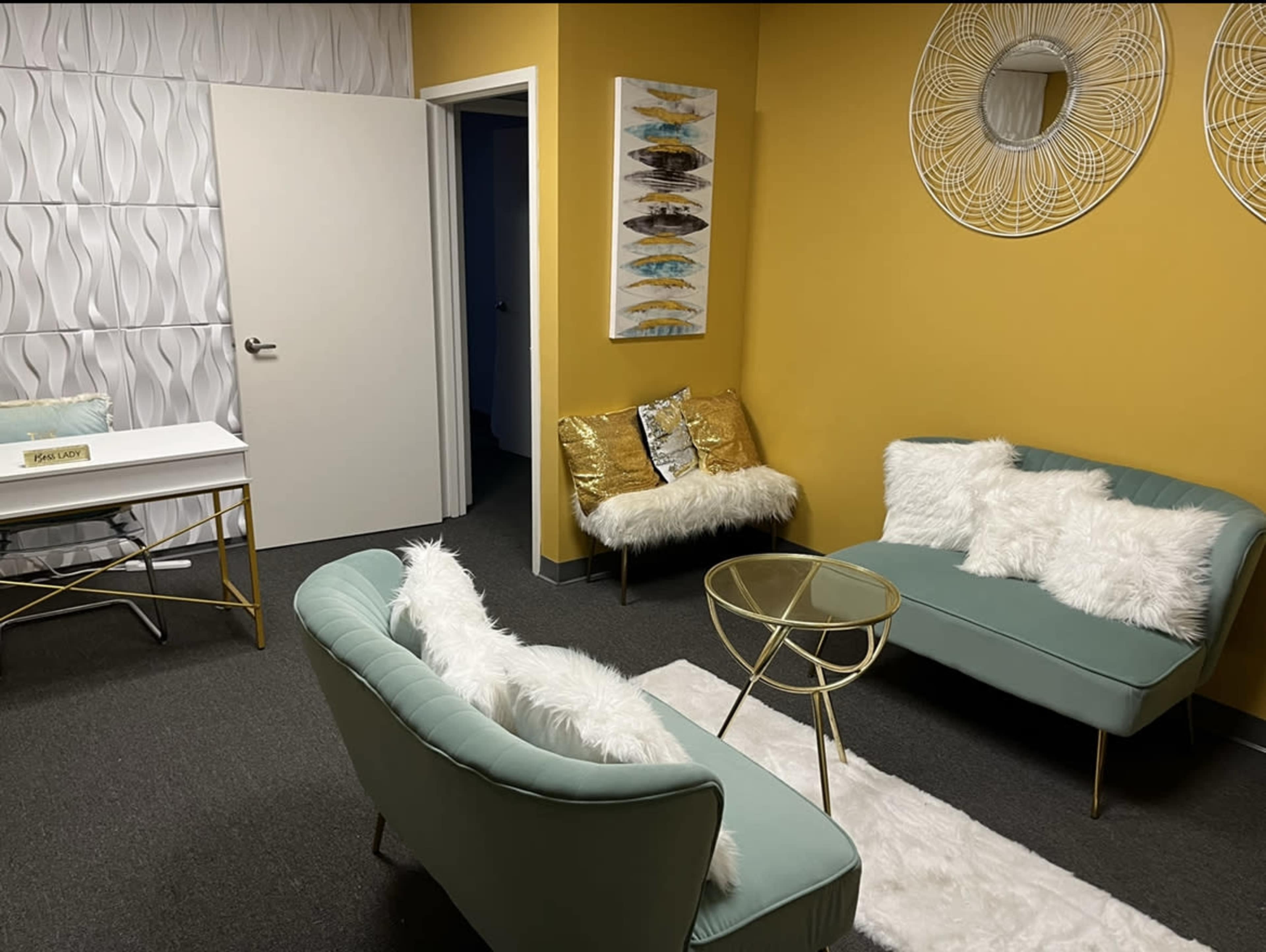 A small, modern seating area with two light green sofas, a glass coffee table, a white rug, and yellow walls, complemented by decorative artwork and mirrors.
