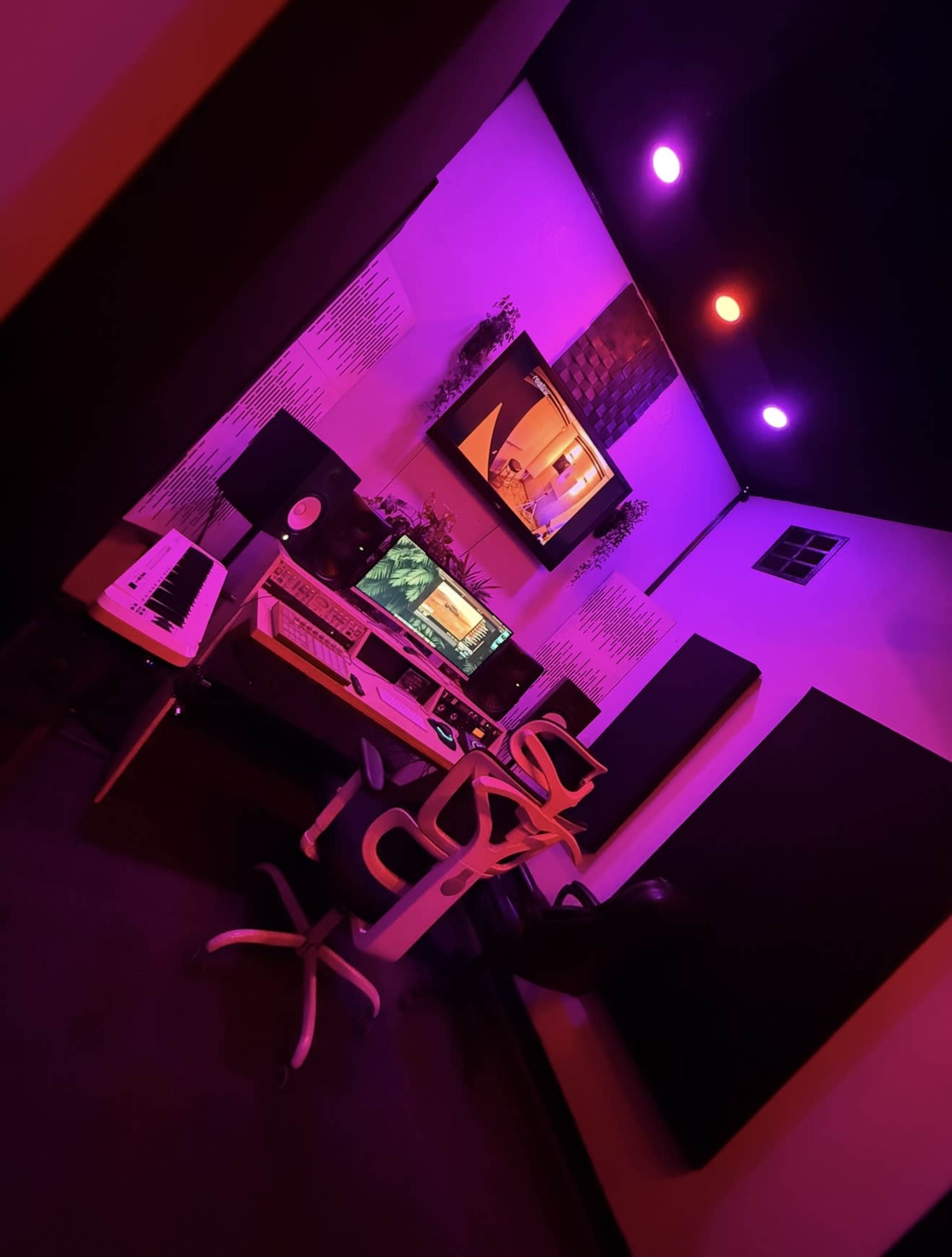 The image shows a colorful music studio with purple and red lighting, featuring a desk with music production equipment and two chairs.