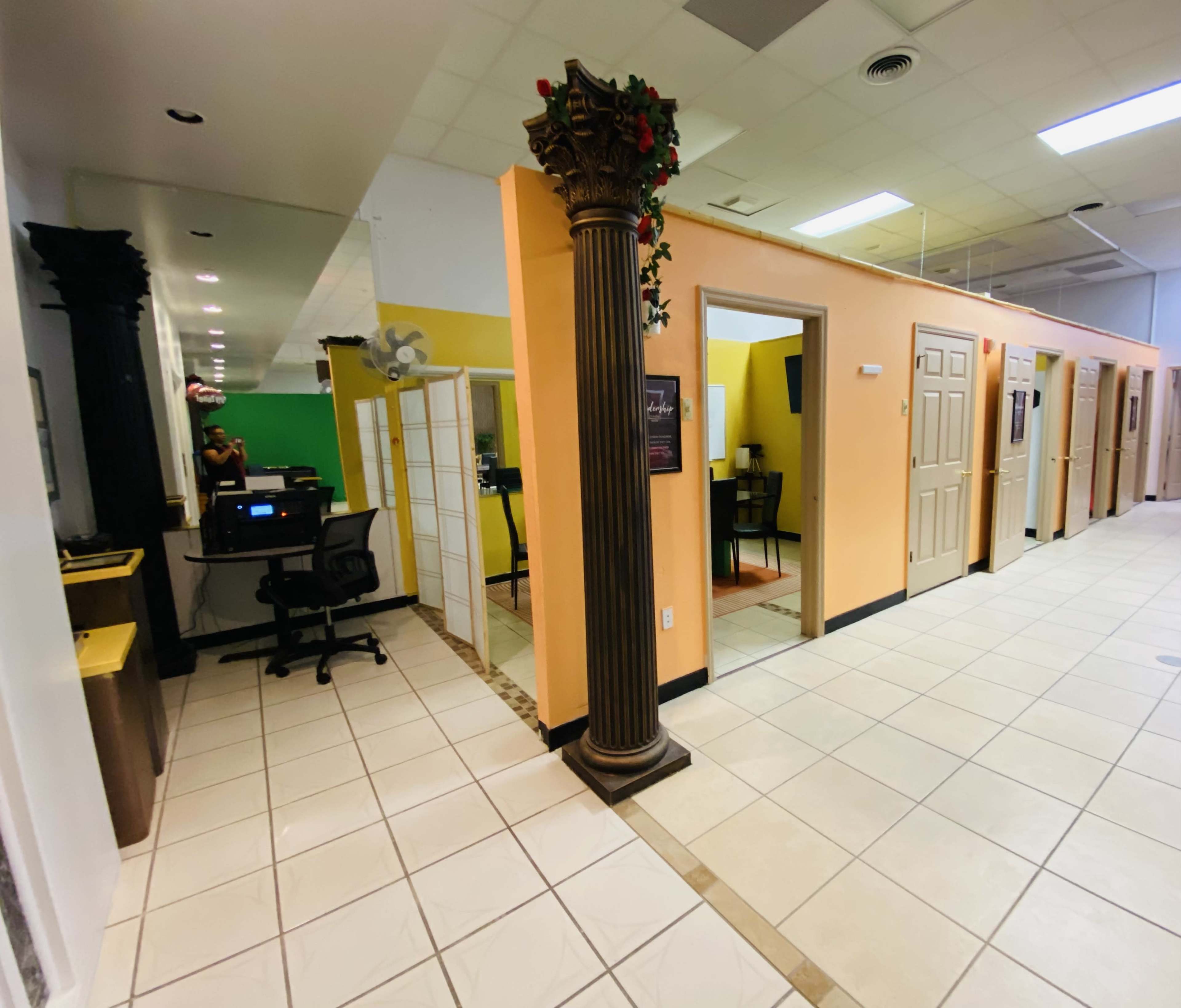 A hallway with several closed doors on the right and an office area with a computer on the left, framed by a decorative column.