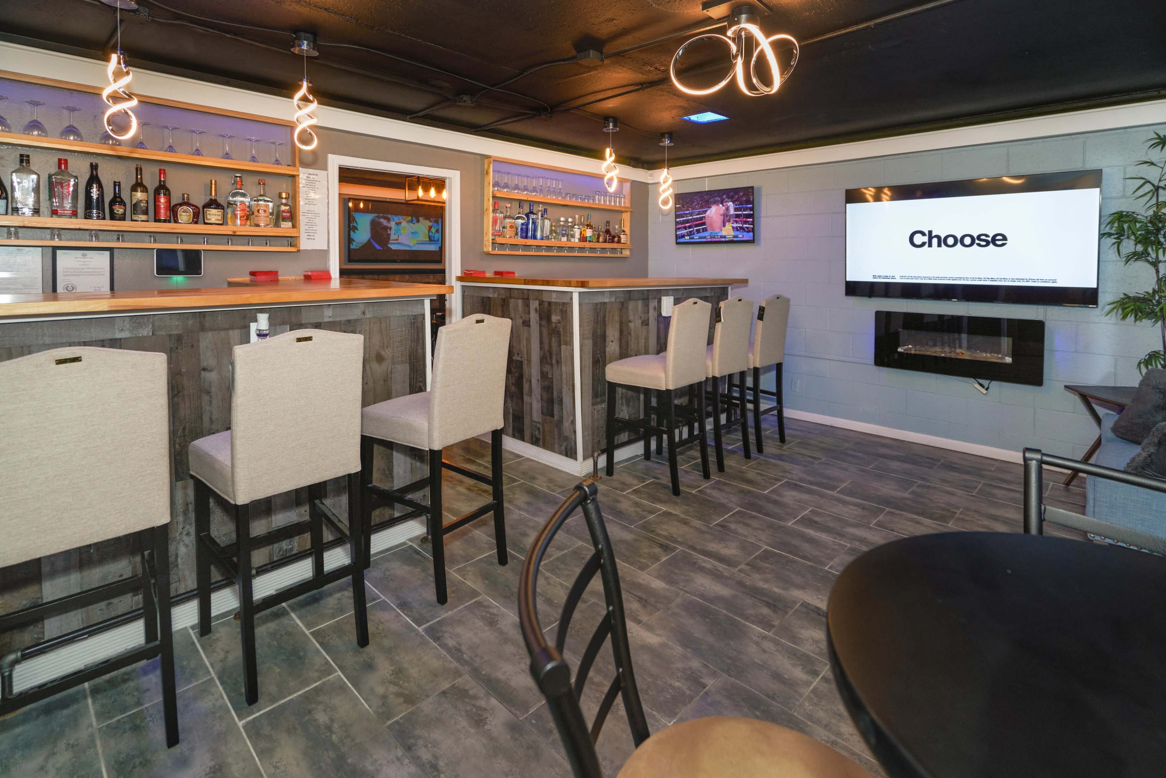 A modern bar area with high stools, shelves stocked with various bottles, and a large screen displaying the word "Choose."