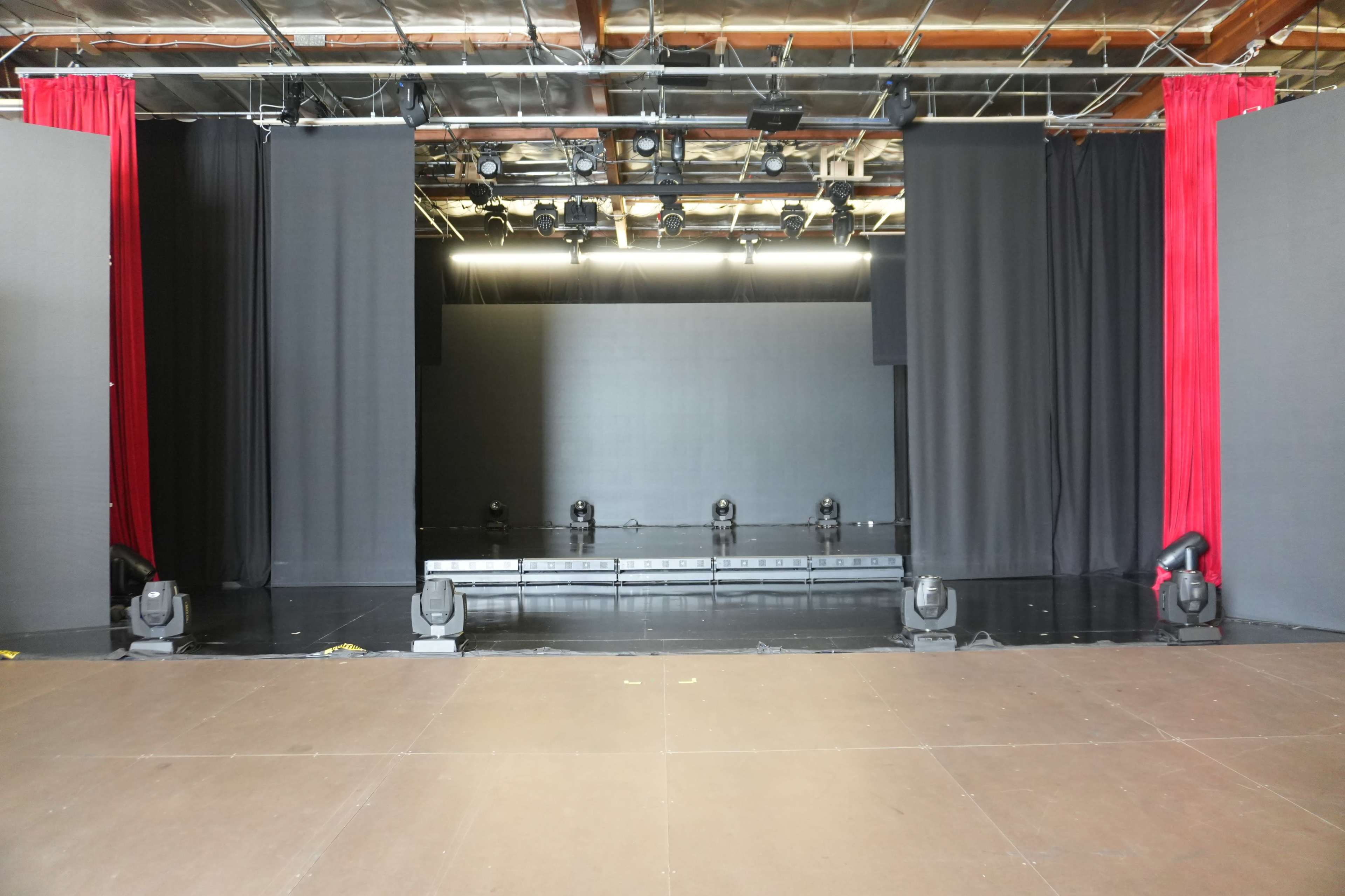The image shows an empty stage with black curtains and red drapes, illuminated by overhead lights.