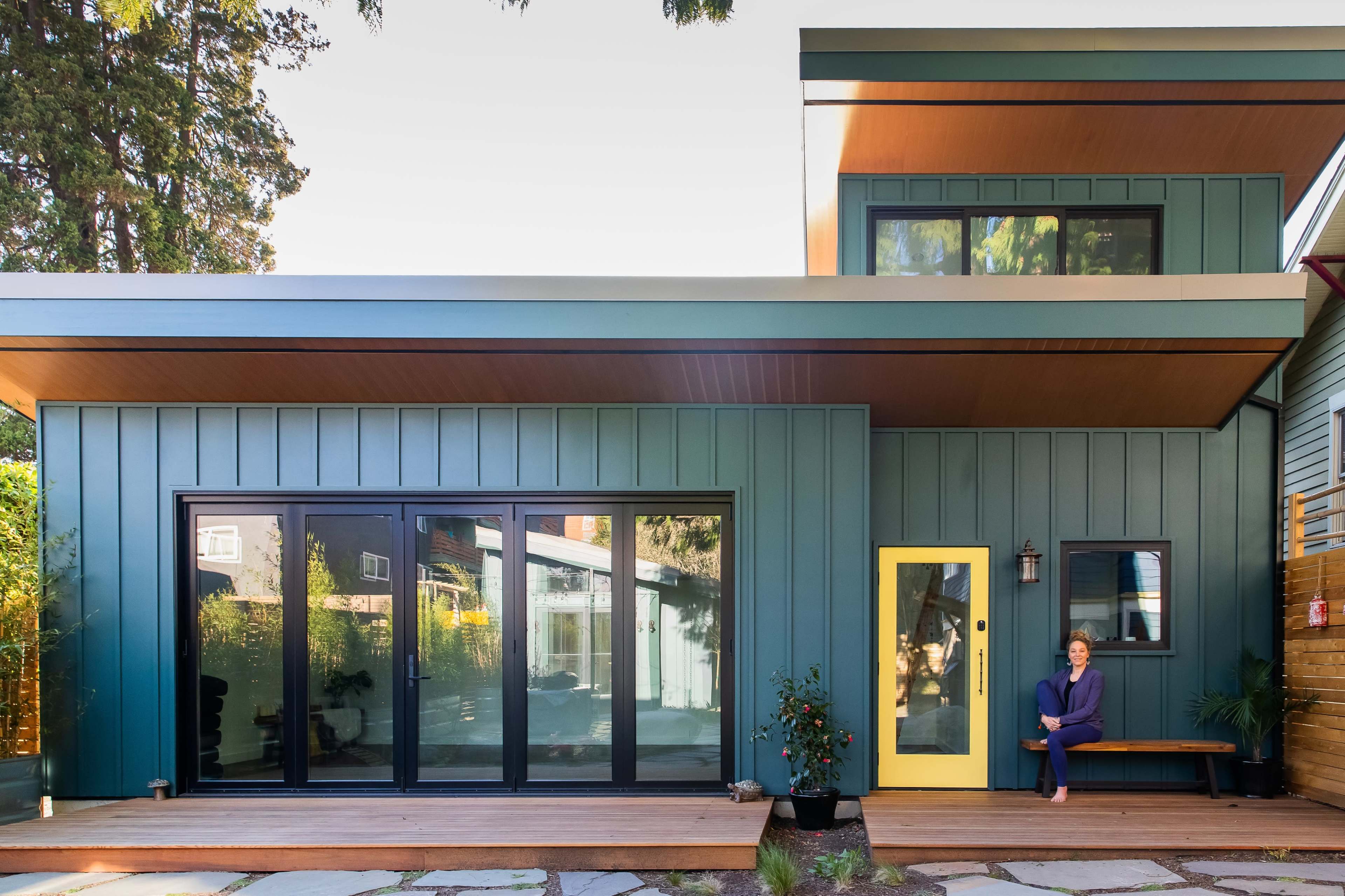 A contemporary house with dark blue siding, large glass doors, and a bright yellow front door features a person sitting on a wooden bench.