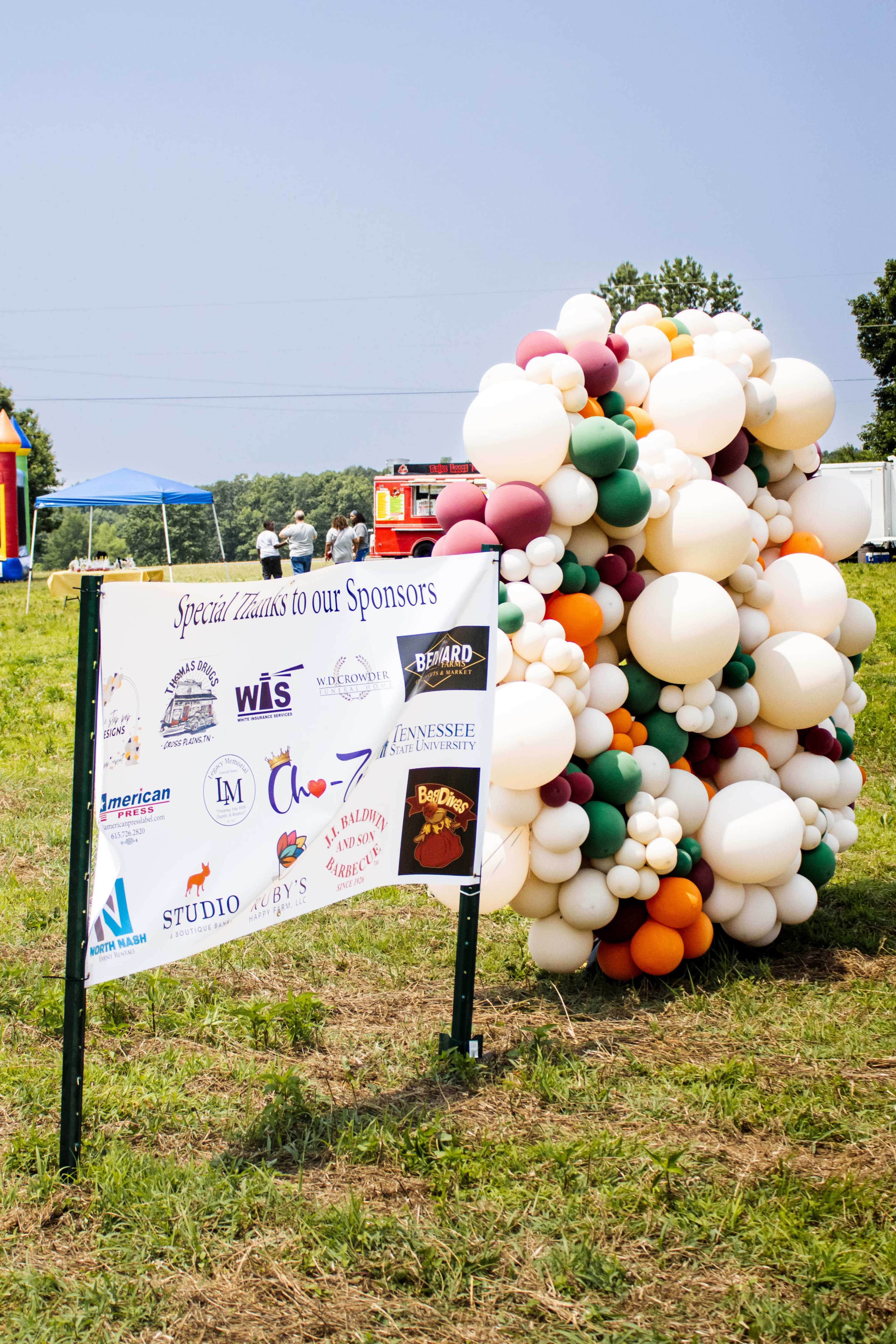 A colorful balloon arrangement stands beside a sign thanking sponsors at an outdoor event.
