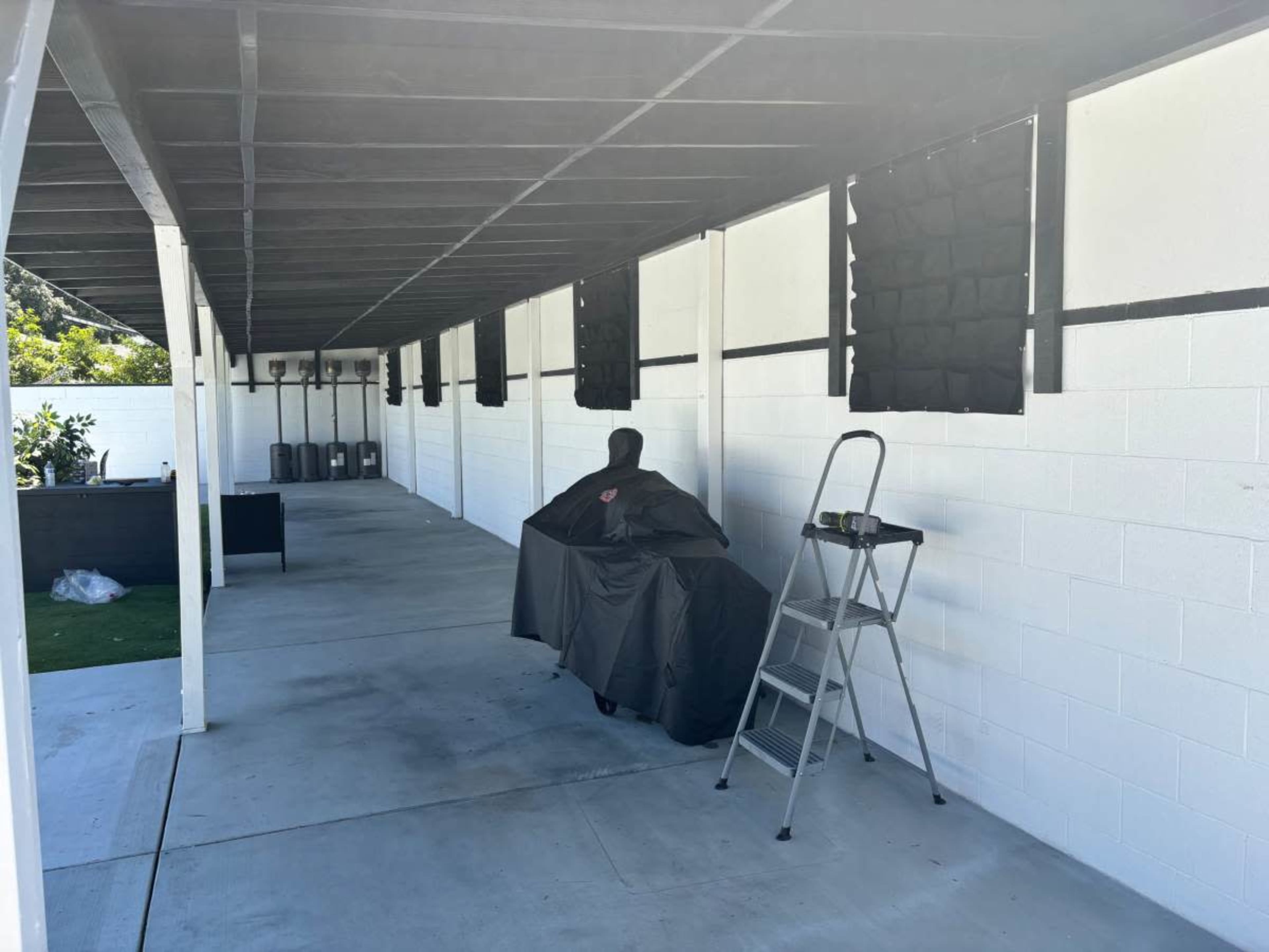 A covered object in a covered patio area with a ladder and several wall-mounted shades.