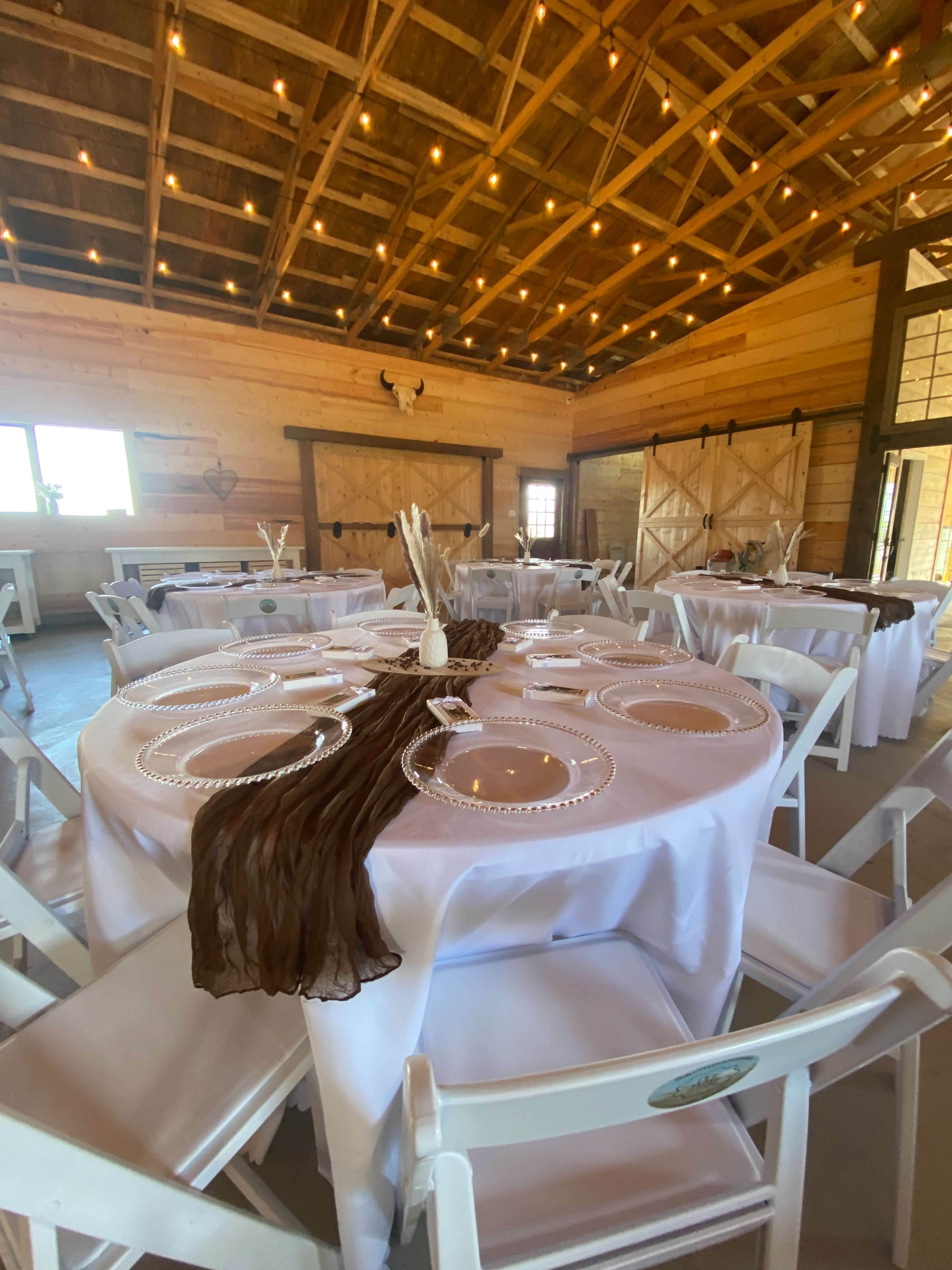 The image shows a decorated indoor event space featuring round tables set with white tablecloths, clear plates, and centerpieces, under warm string lights and wooden beams.
