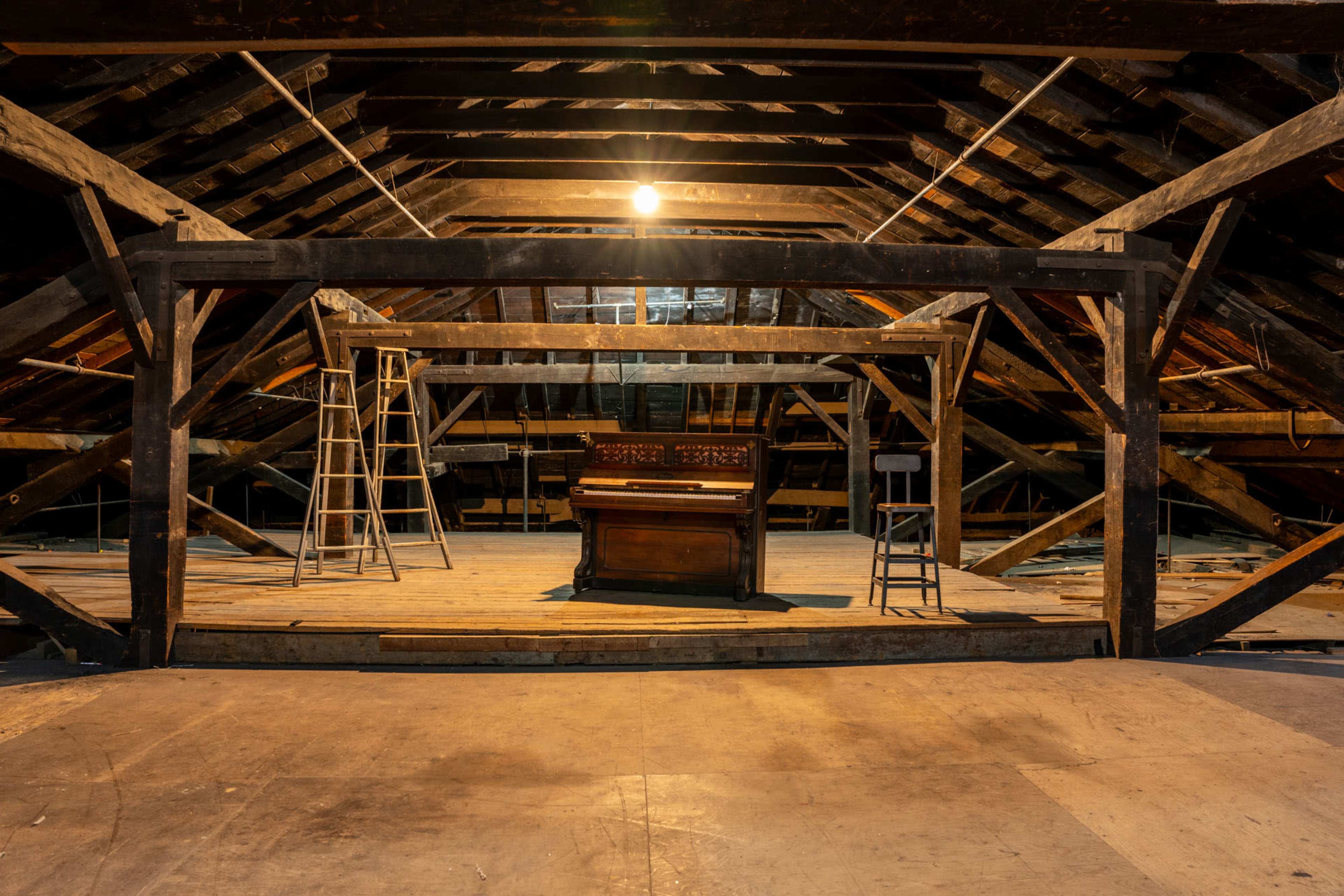 An antique piano is positioned in the center of a spacious, unfinished attic illuminated by a single light.