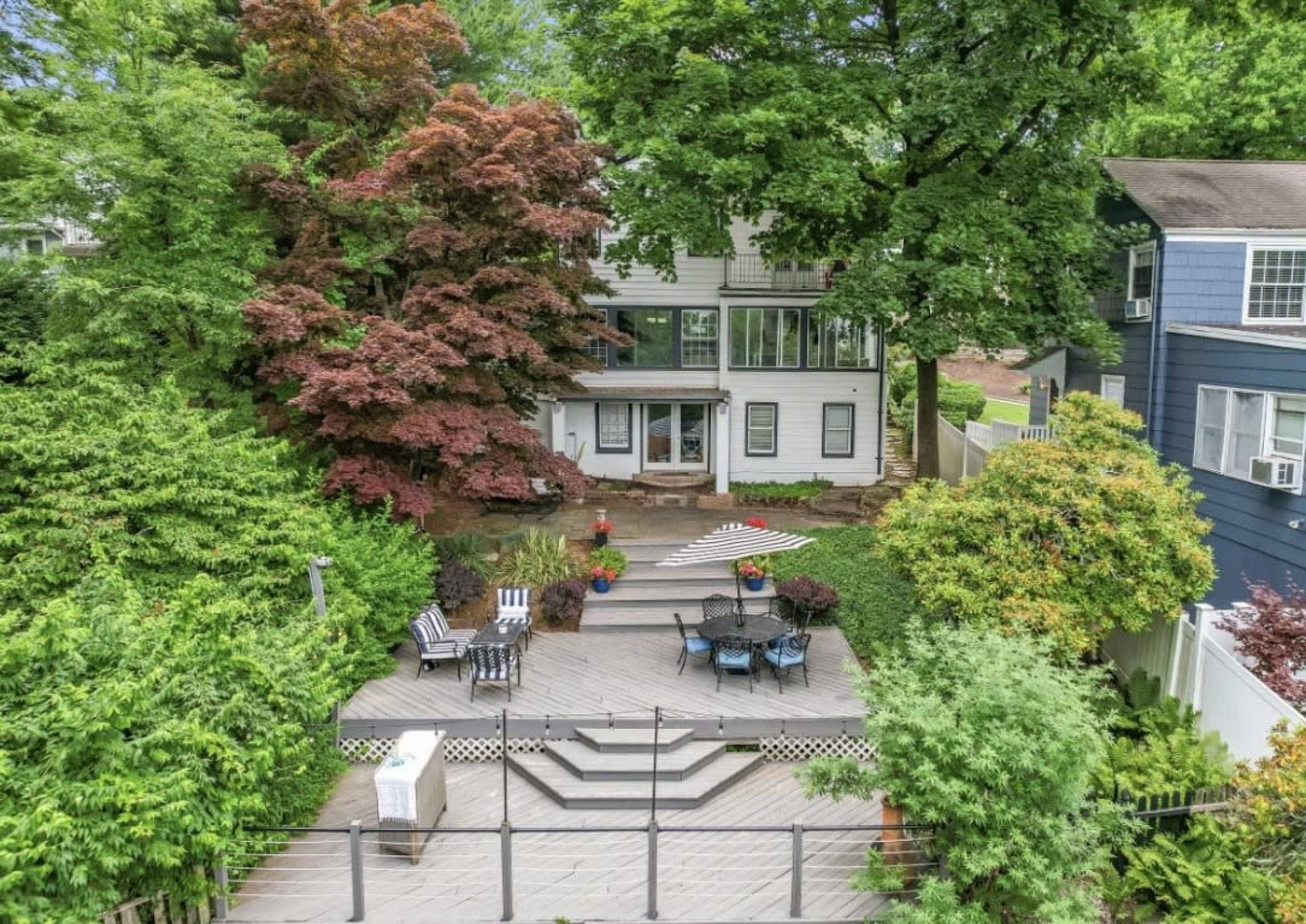 A spacious back yard features a multi-level deck with seating areas, surrounded by lush greenery and trees, in front of a two-story house with large windows.