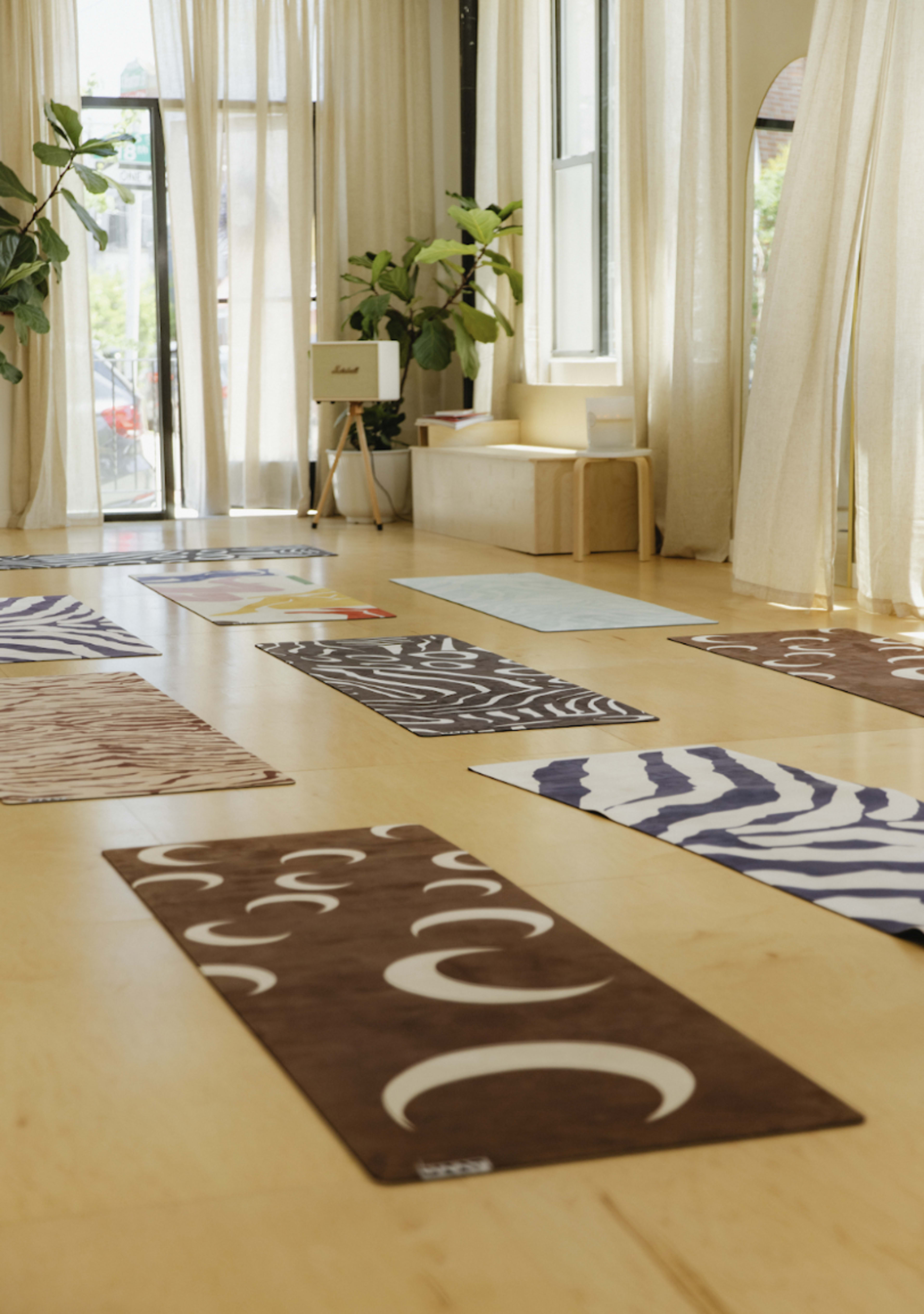 The image shows a bright yoga studio with several patterned mats laid out on a wooden floor and large windows letting in natural light.