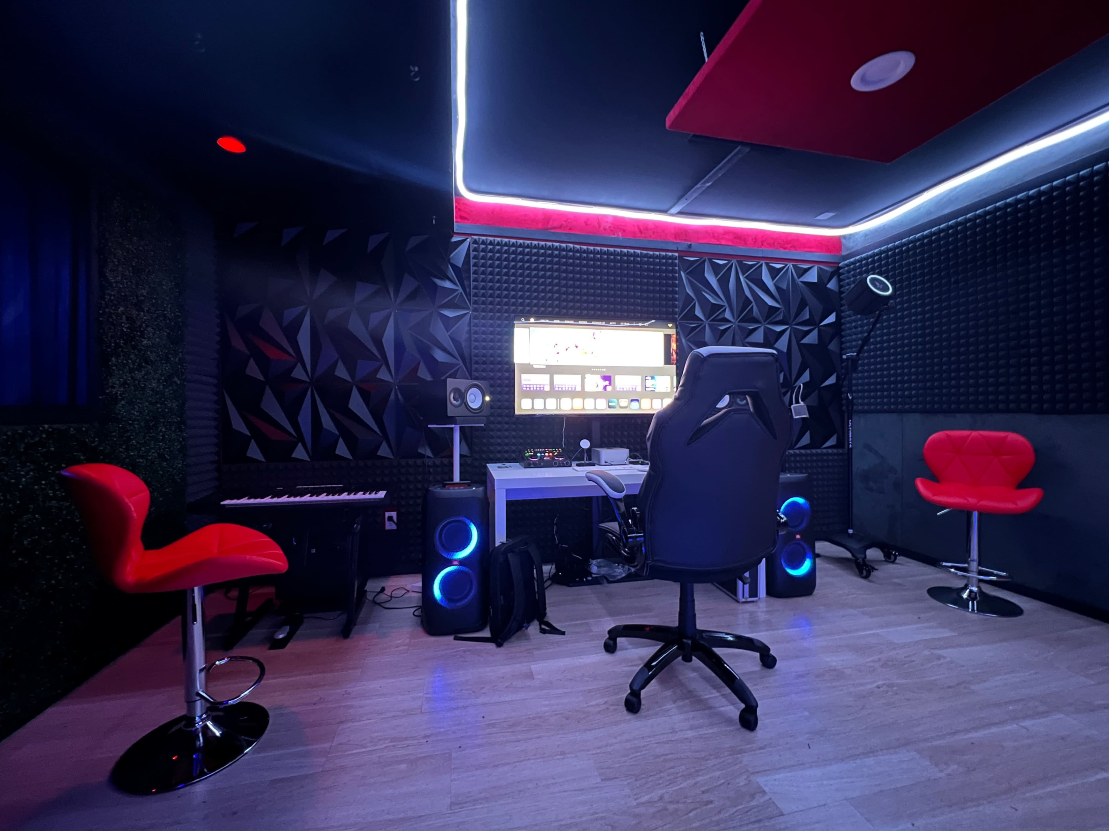 A modern audio studio features a keyboard, a computer setup surrounded by soundproofing panels, and red bar stools.