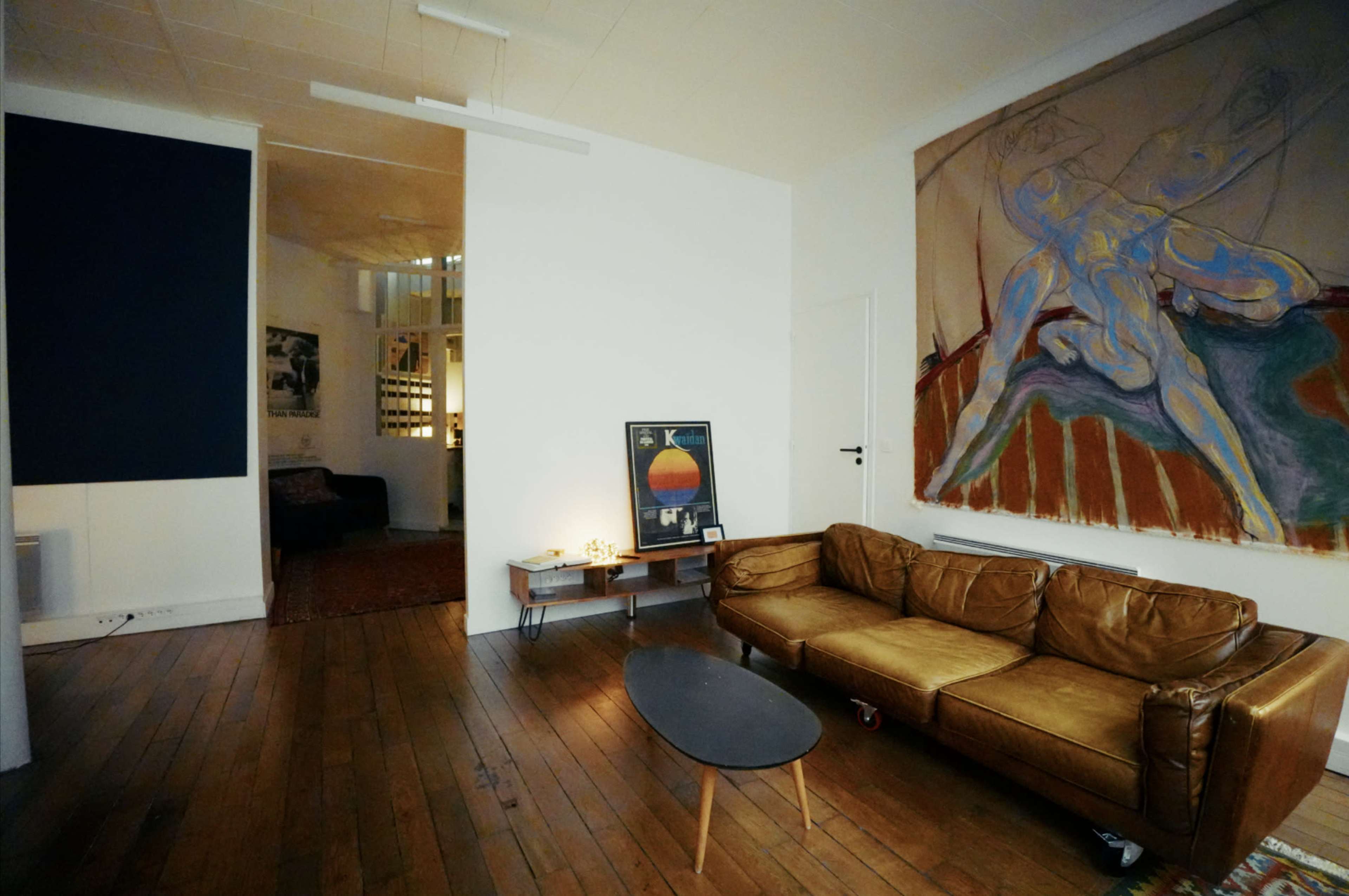 The image shows a modern living room with a brown leather couch, a round coffee table, and a large abstract painting on the wall, with an open doorway leading to a separate area.