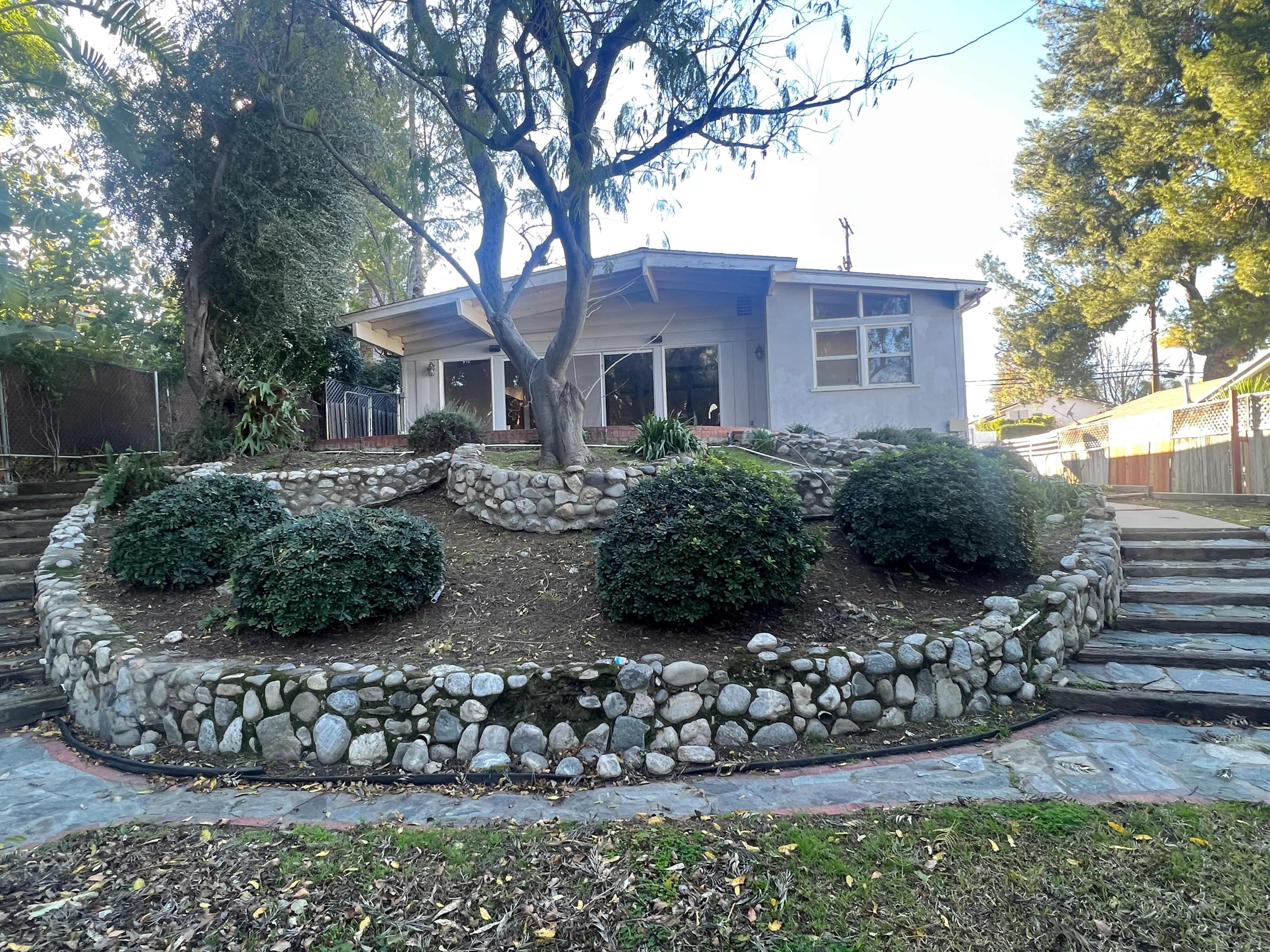 A house with a stone garden featuring circular shrubbery, surrounded by steps and trees.