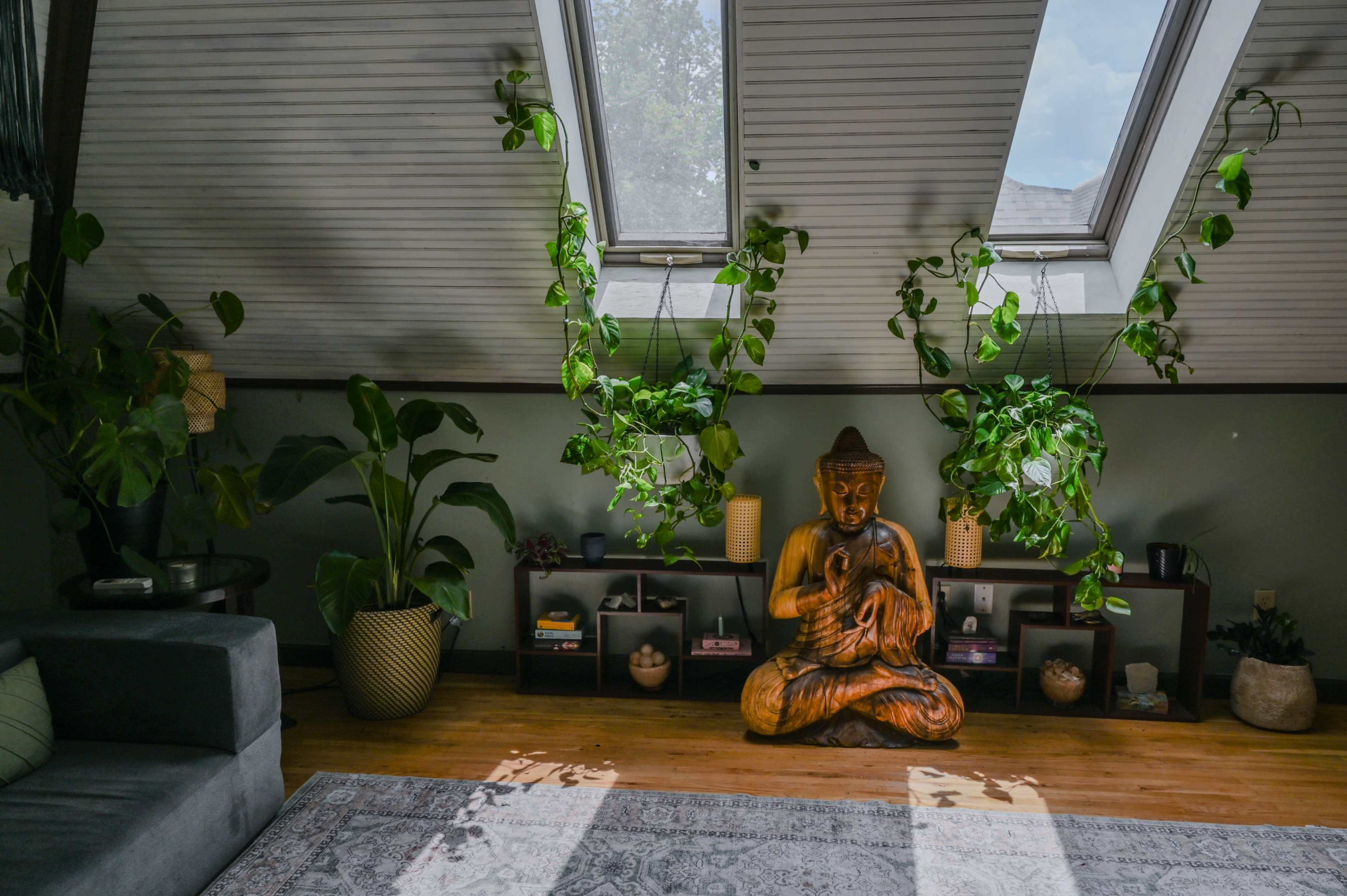 The image depicts a cozy room with a wooden Buddha sculpture surrounded by plants and shelves, illuminated by natural light coming through skylights.