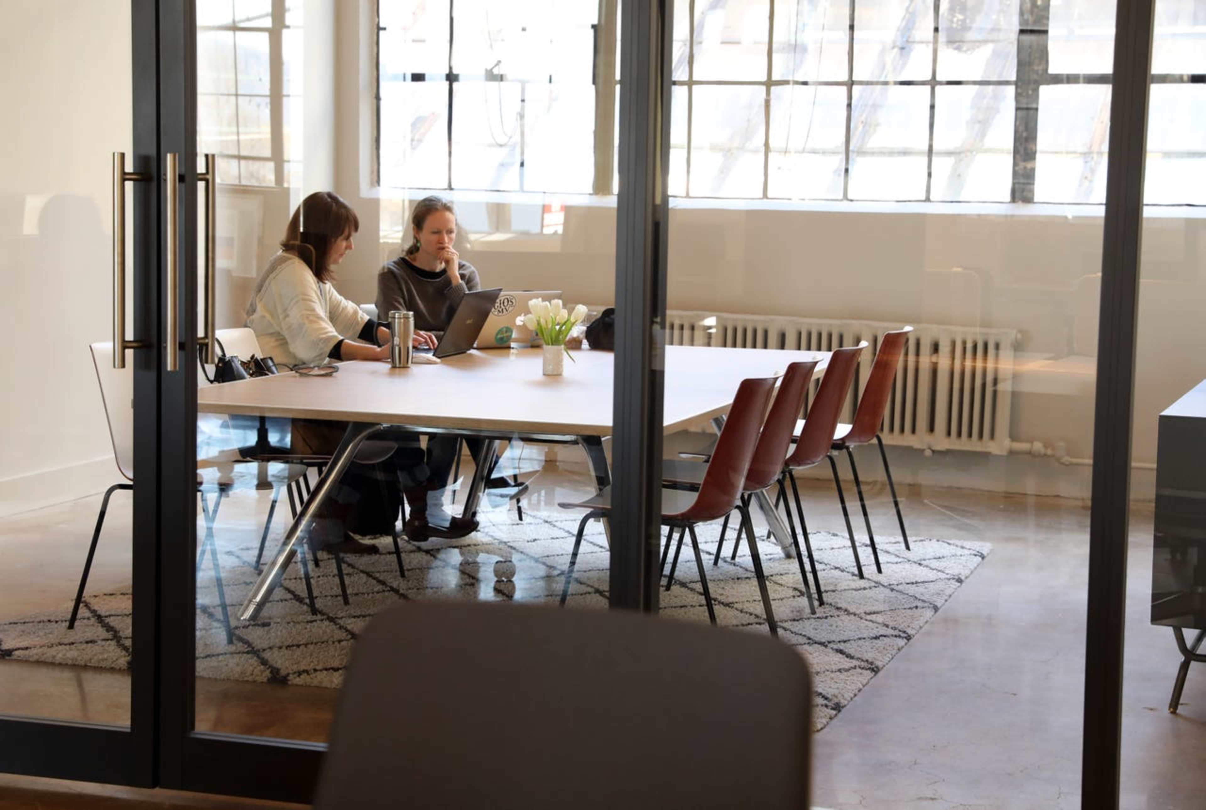 Two women are sitting at a table in a well-lit office, working on laptops with a vase of flowers in the center.