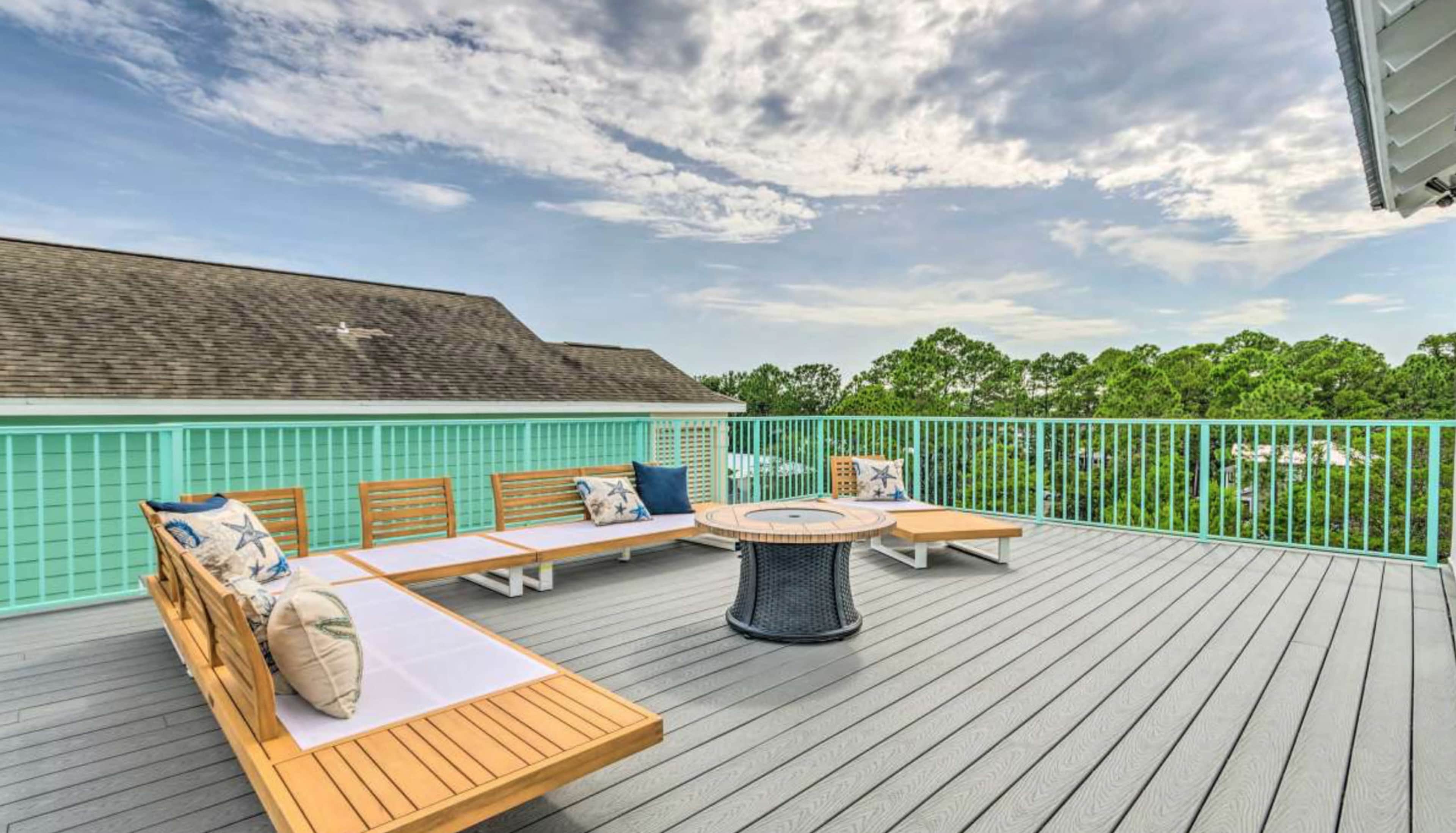 A spacious outdoor deck with wooden seating around a circular table, surrounded by green trees under a partly cloudy sky.