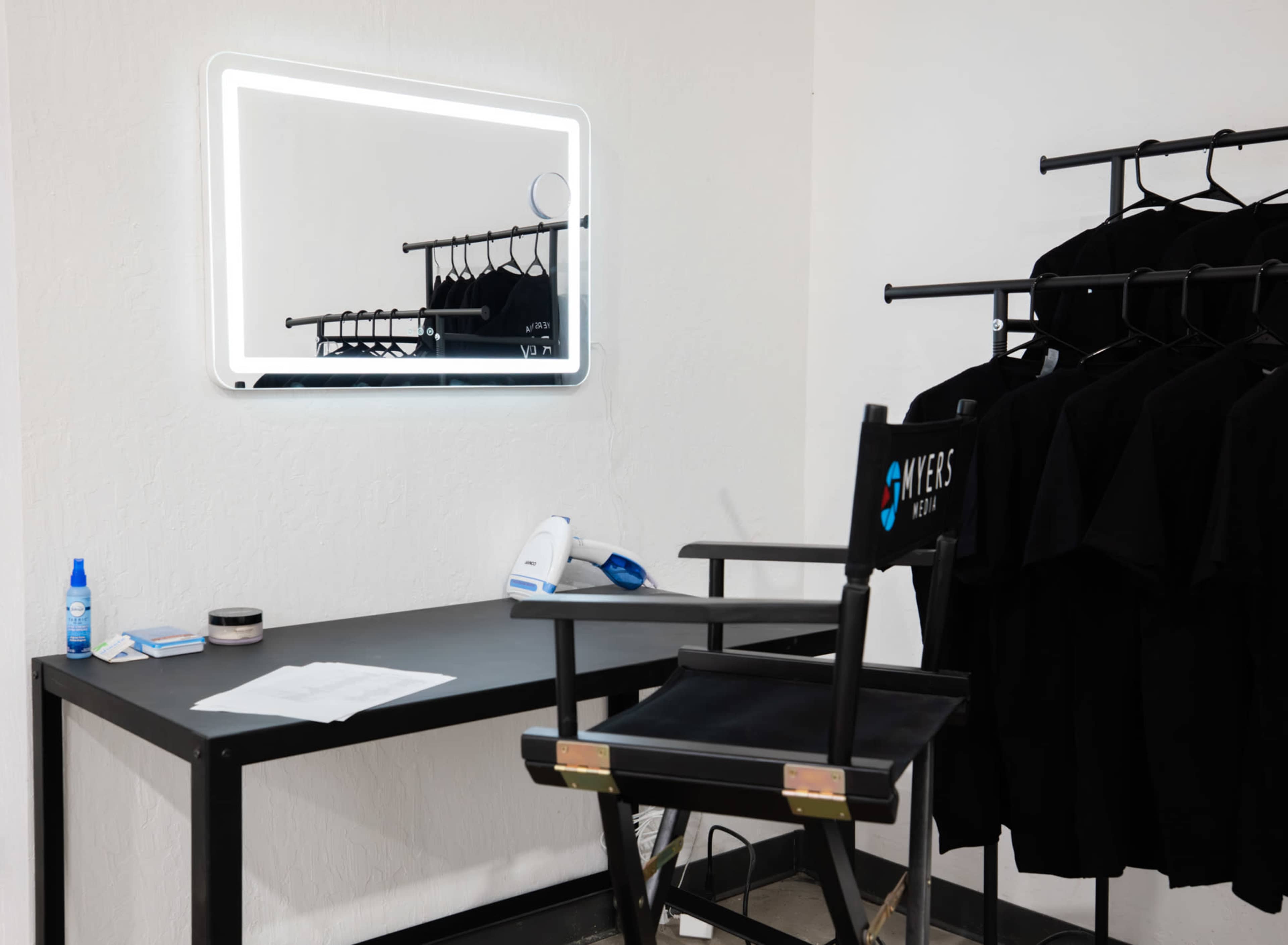 The image shows a neatly arranged makeup station featuring a mirror with backlighting, a chair, a desk with some tools, and several black clothing items hanging on a rack.