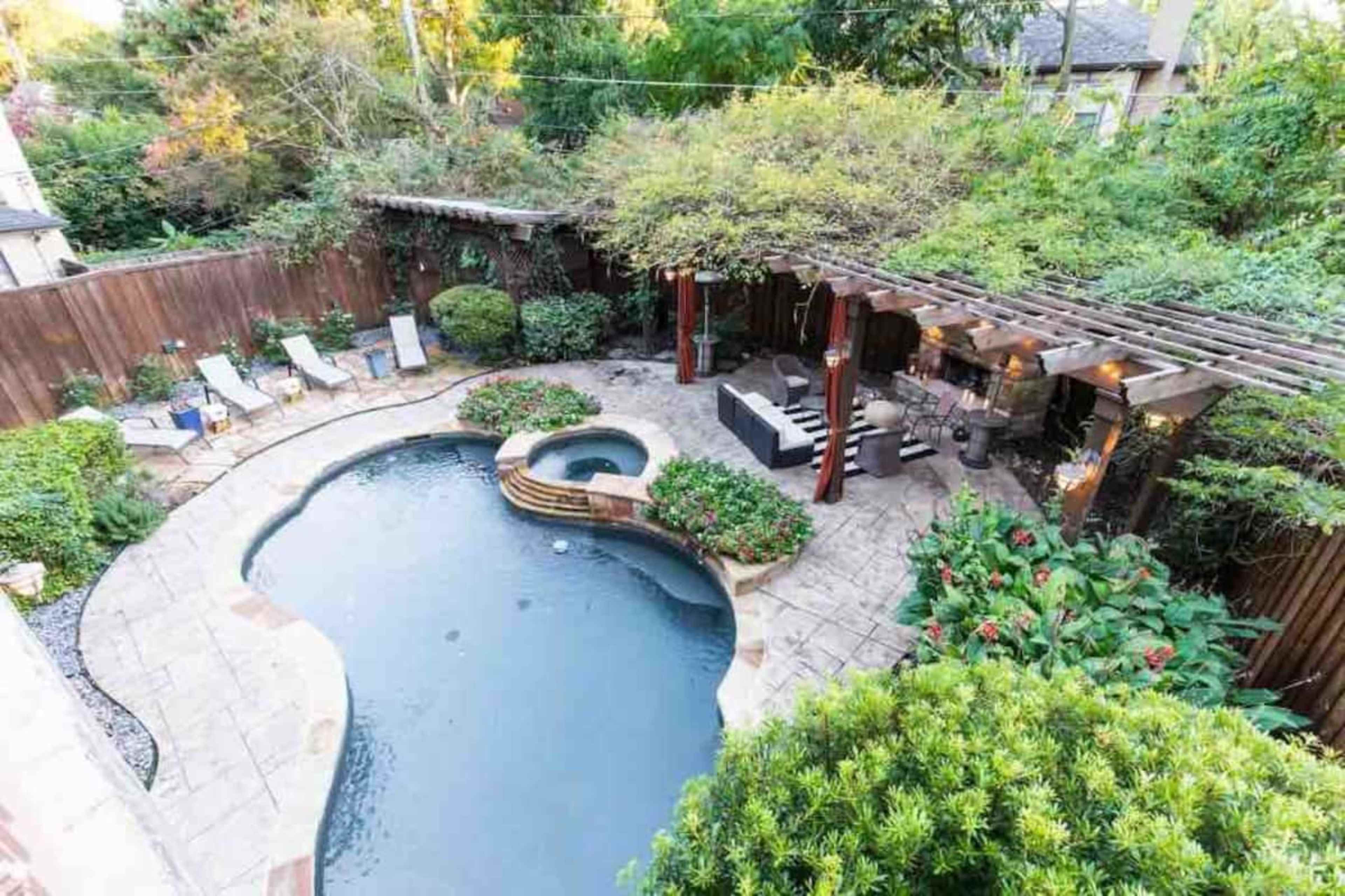 The image shows a backyard with a swimming pool, a spa area, stone patio, lounge chairs, and a shaded pergola surrounded by lush greenery.