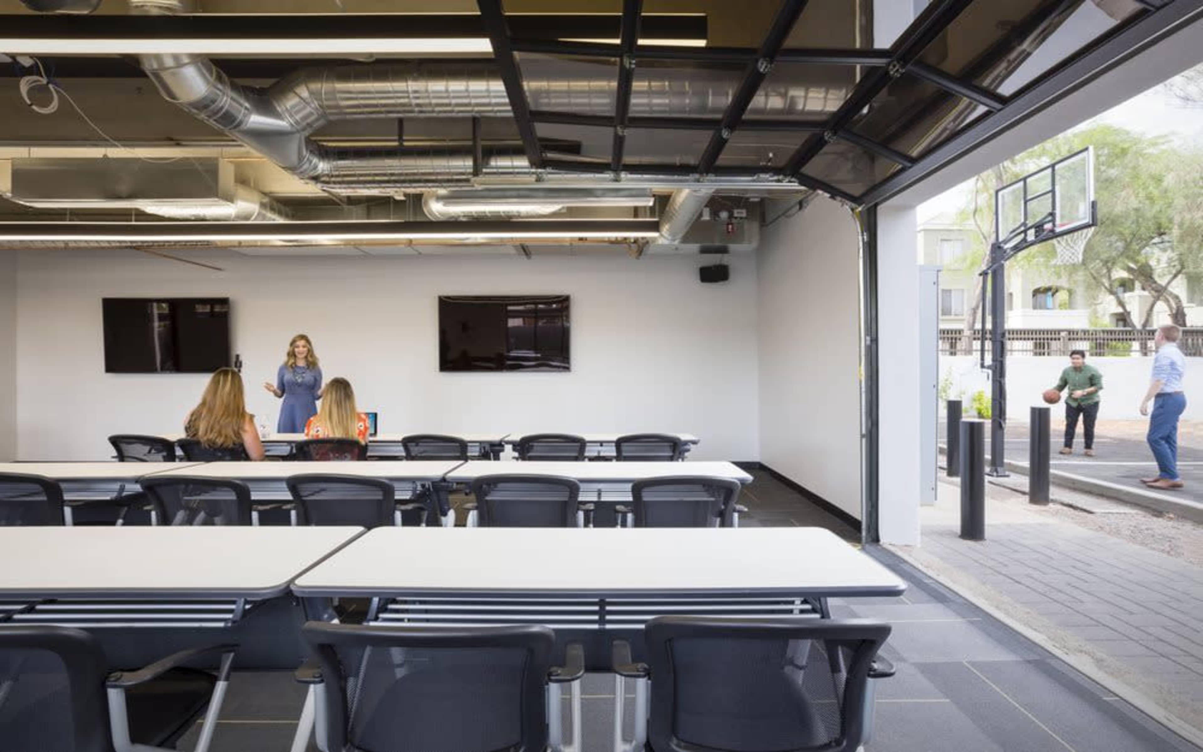 A modern conference room features several tables facing a screen, with a woman presenting to a small audience, while two people play basketball outside through an open garage door.