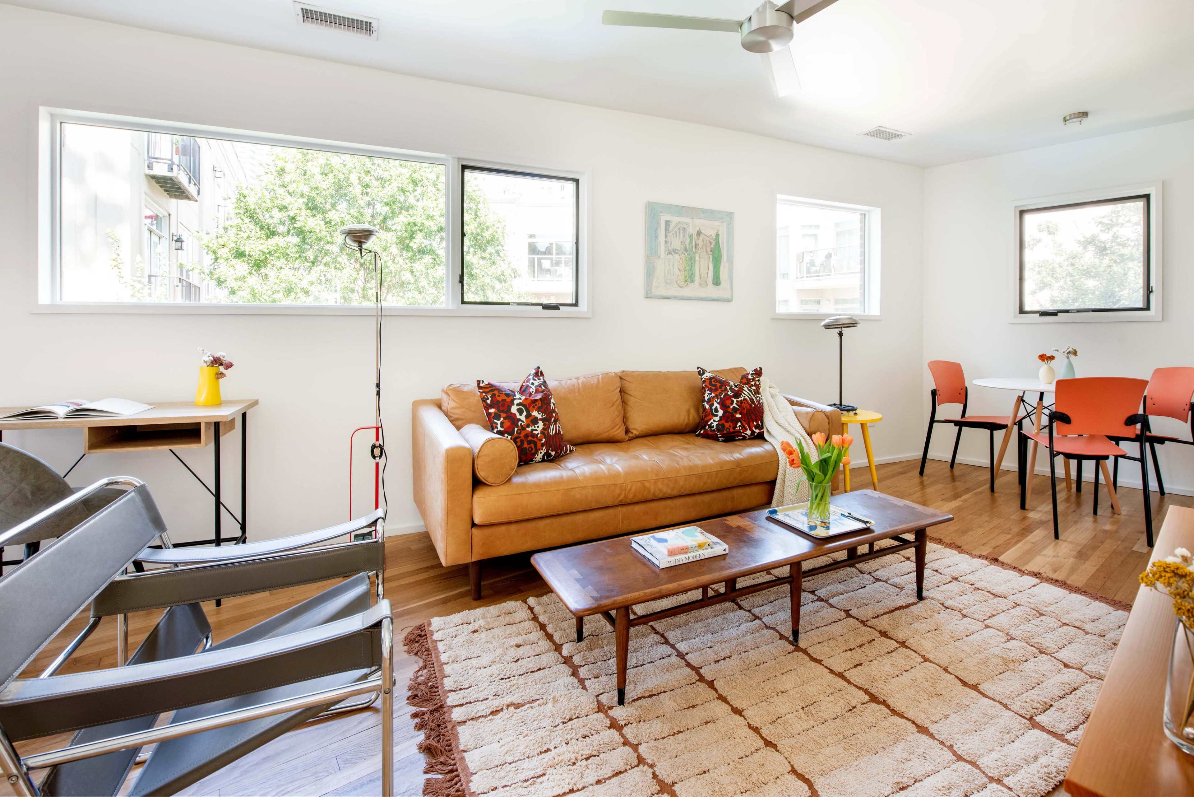 The image features a well-lit living area with a brown leather sofa, a coffee table, and a small dining area with orange chairs.