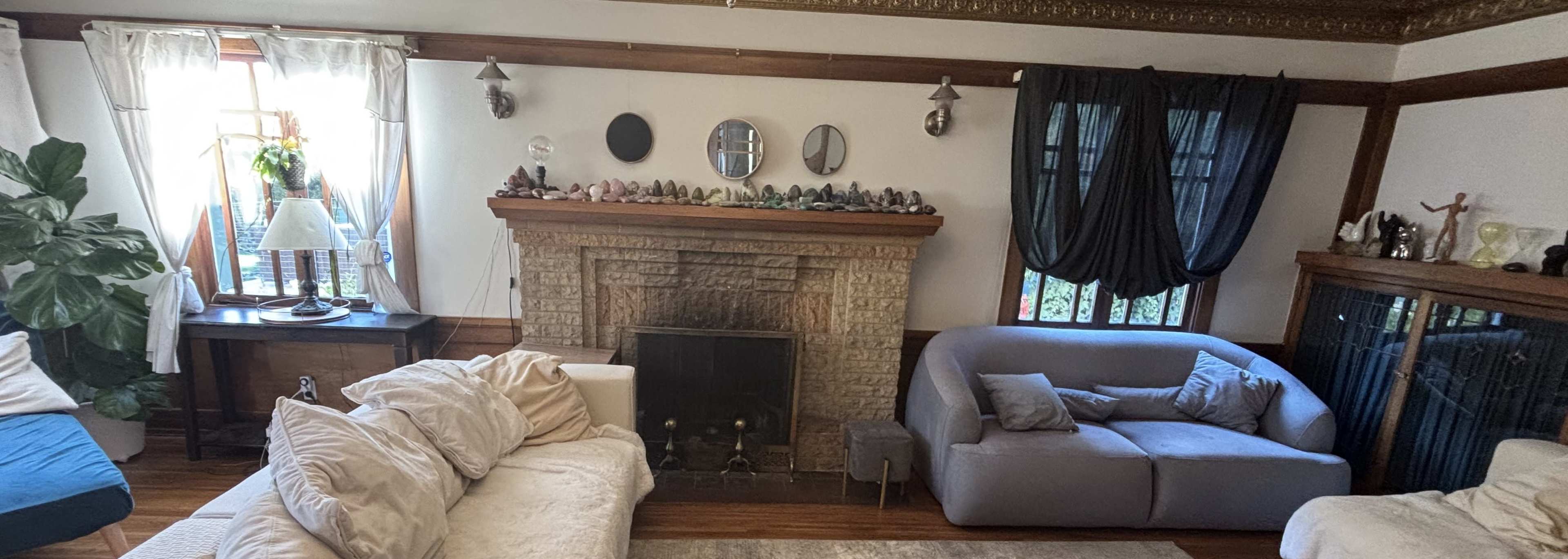 The image shows a cozy living room with two sofas, a stone fireplace, and a window adorned with decorative plants and crystals.