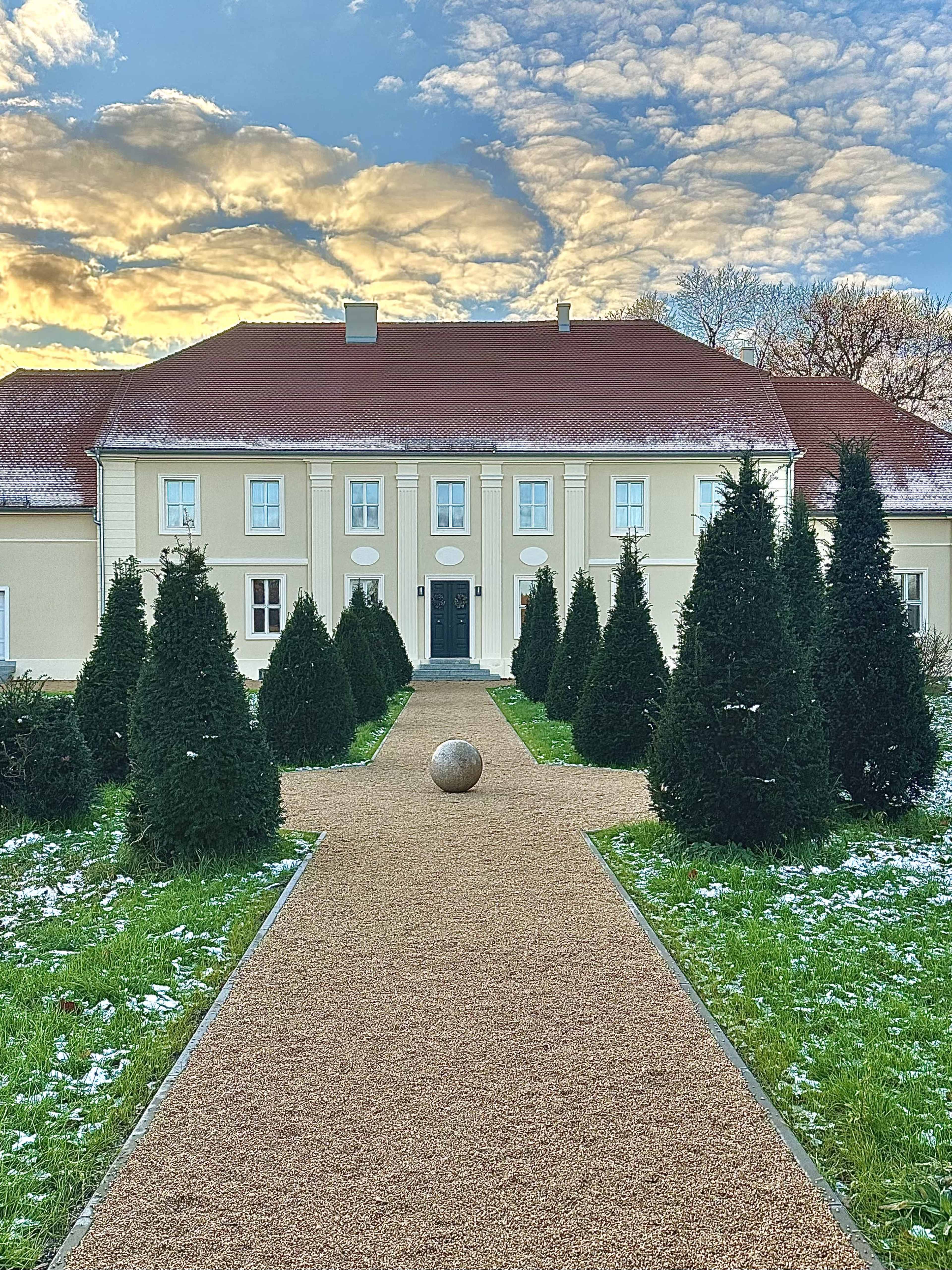 A gravel path leads to a stately building with a red roof, flanked by conical evergreen trees and a spherical sculpture on the ground, under a sky with clouds at sunset.