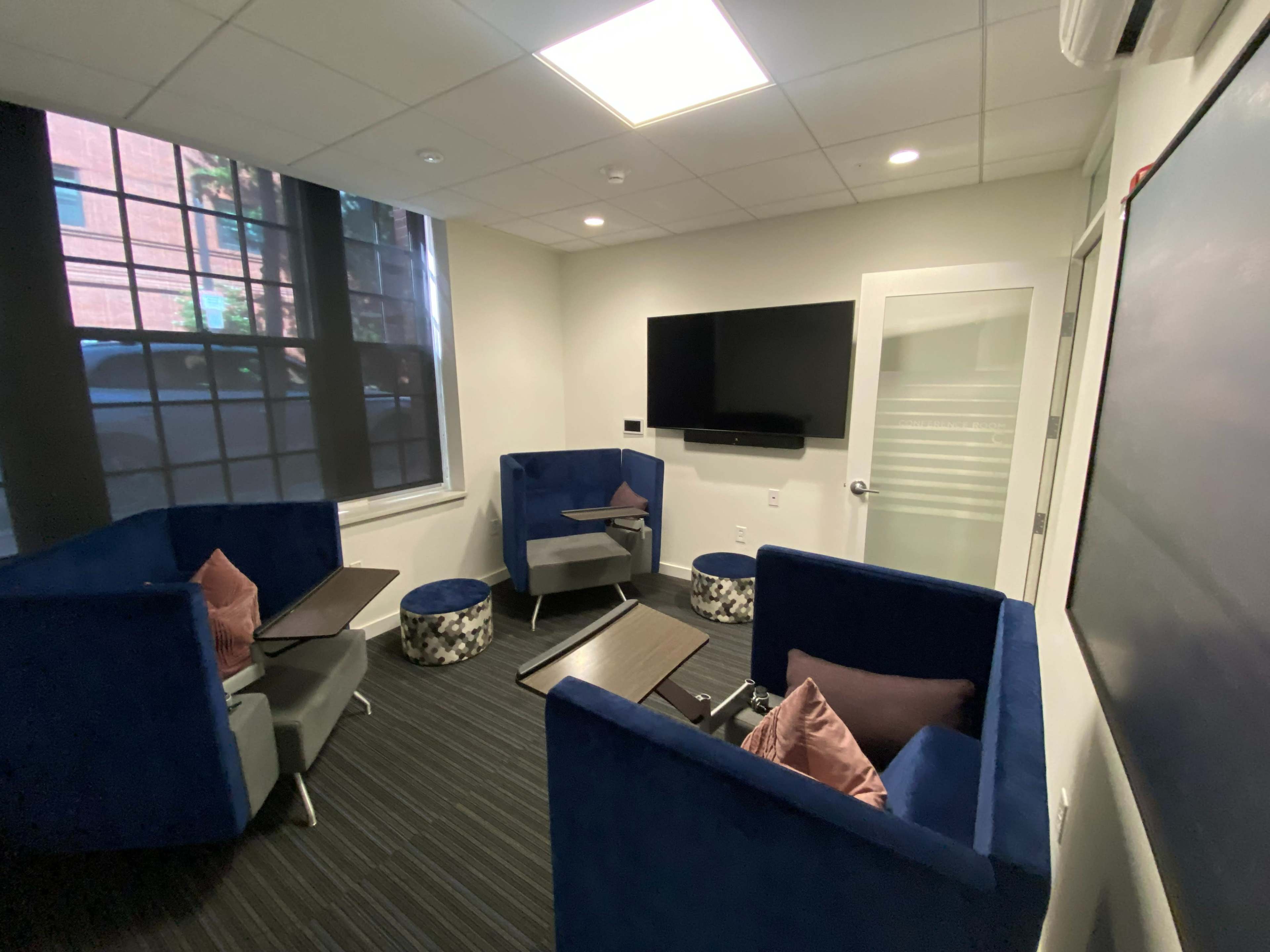 A small meeting room equipped with two blue couches, a wall-mounted television, and a large window.