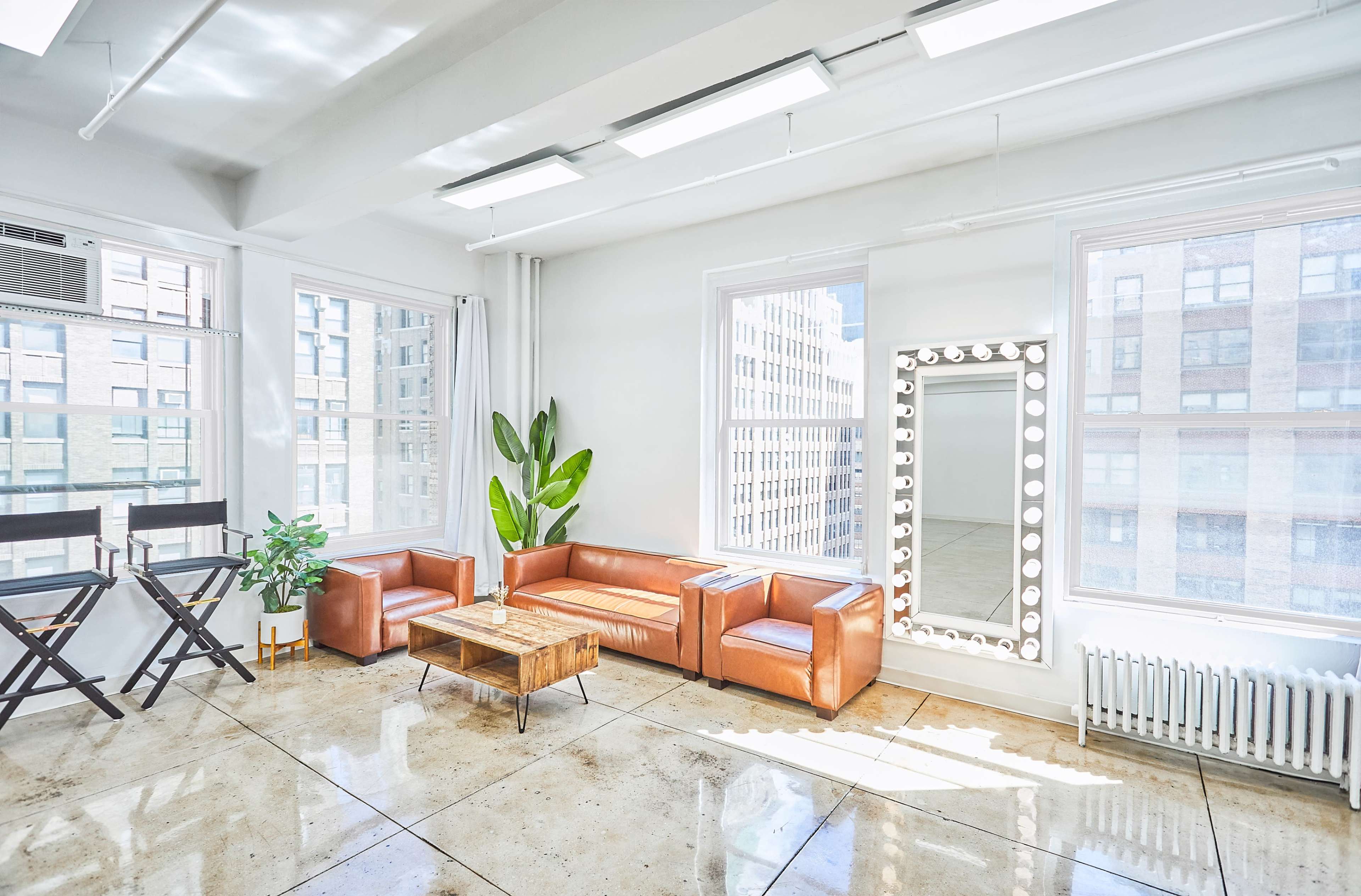 The image shows a bright, modern living space featuring a brown leather couch, a wooden coffee table, a large illuminated mirror, and tall windows with a view of a cityscape.