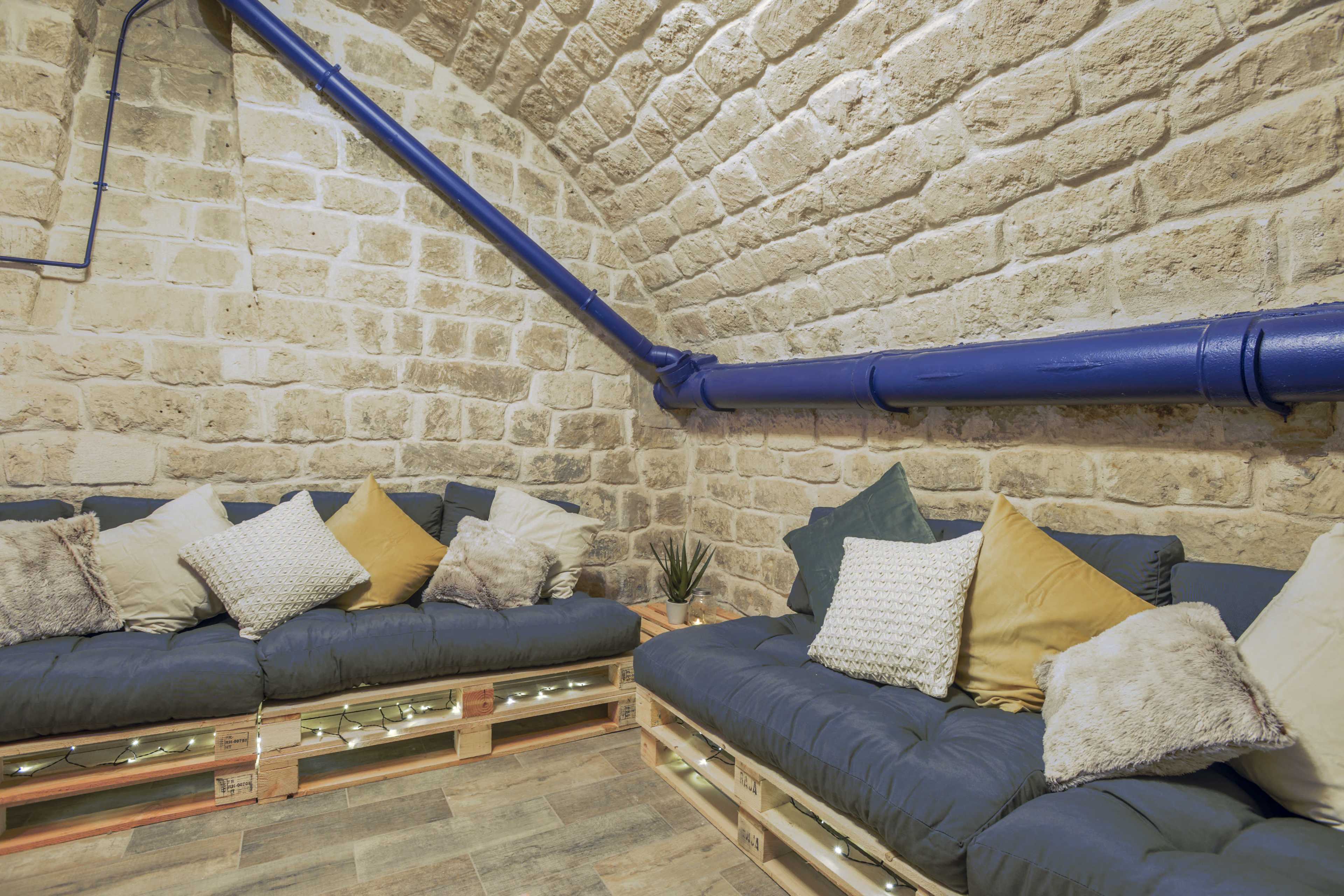 A cozy seating area with pallet couches adorned with various cushions against a stone wall, complemented by a blue pipe running along the ceiling.