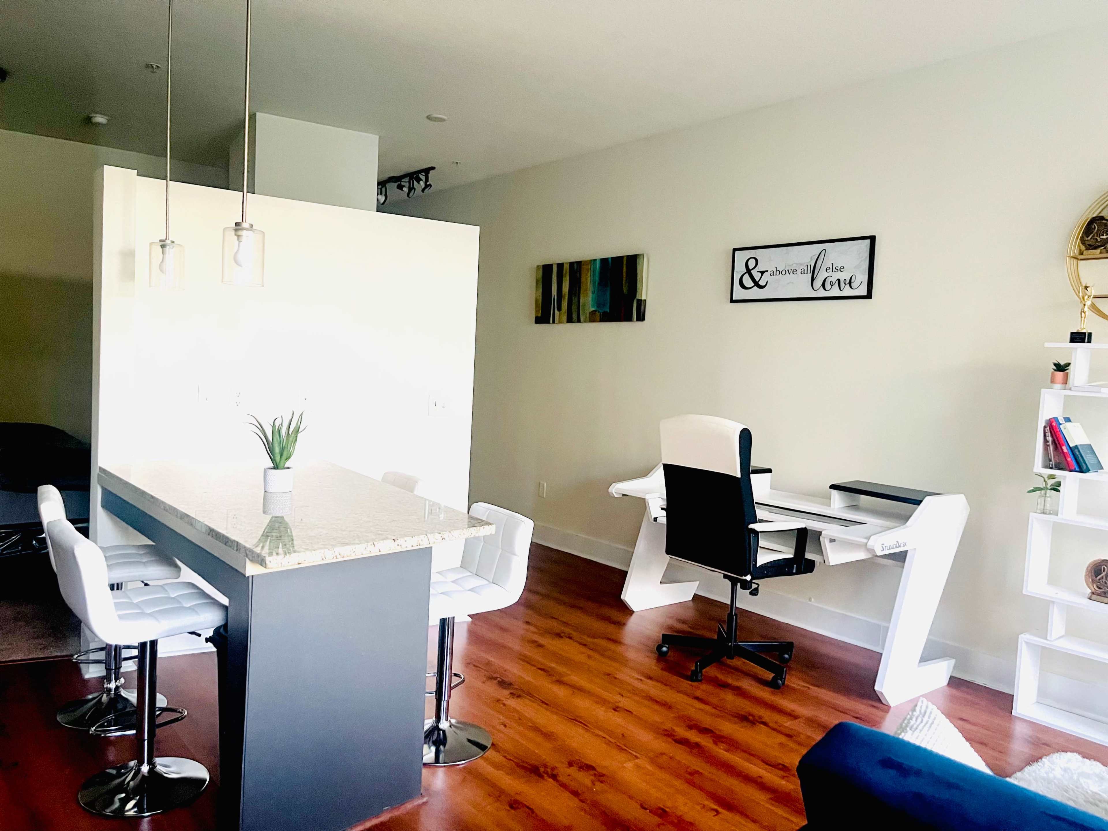 The image shows a modern living space featuring a kitchen island with four white bar stools, a workspace with a white desk and chair, and light wooden flooring.