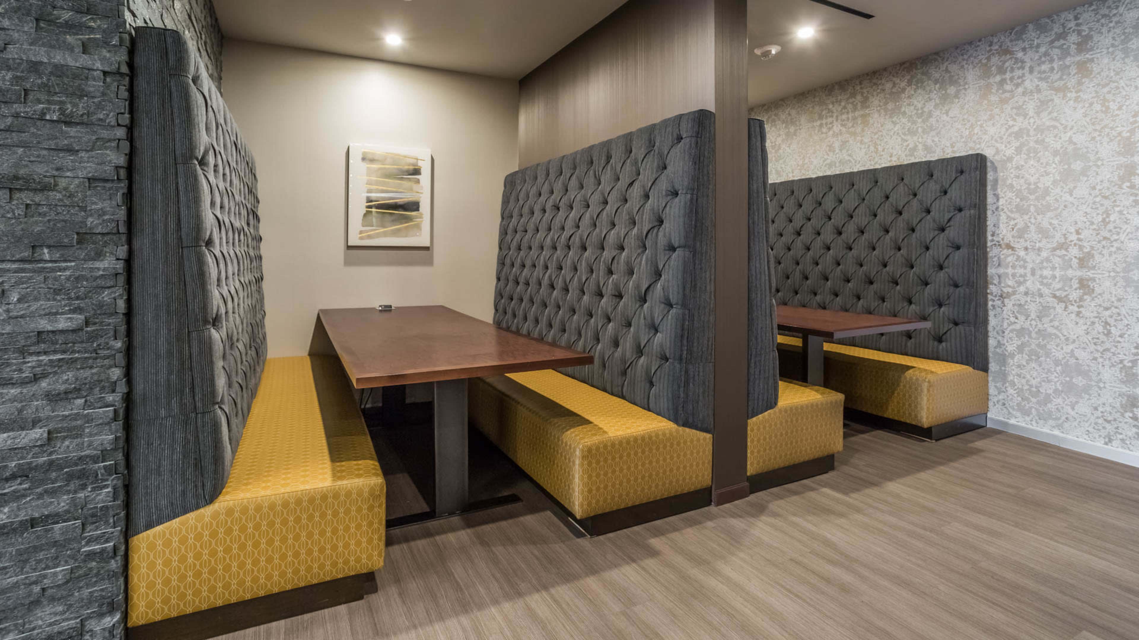 The image shows a modern restaurant interior featuring two tufted booths with wooden tabletops and patterned seating, set against a textured wall.