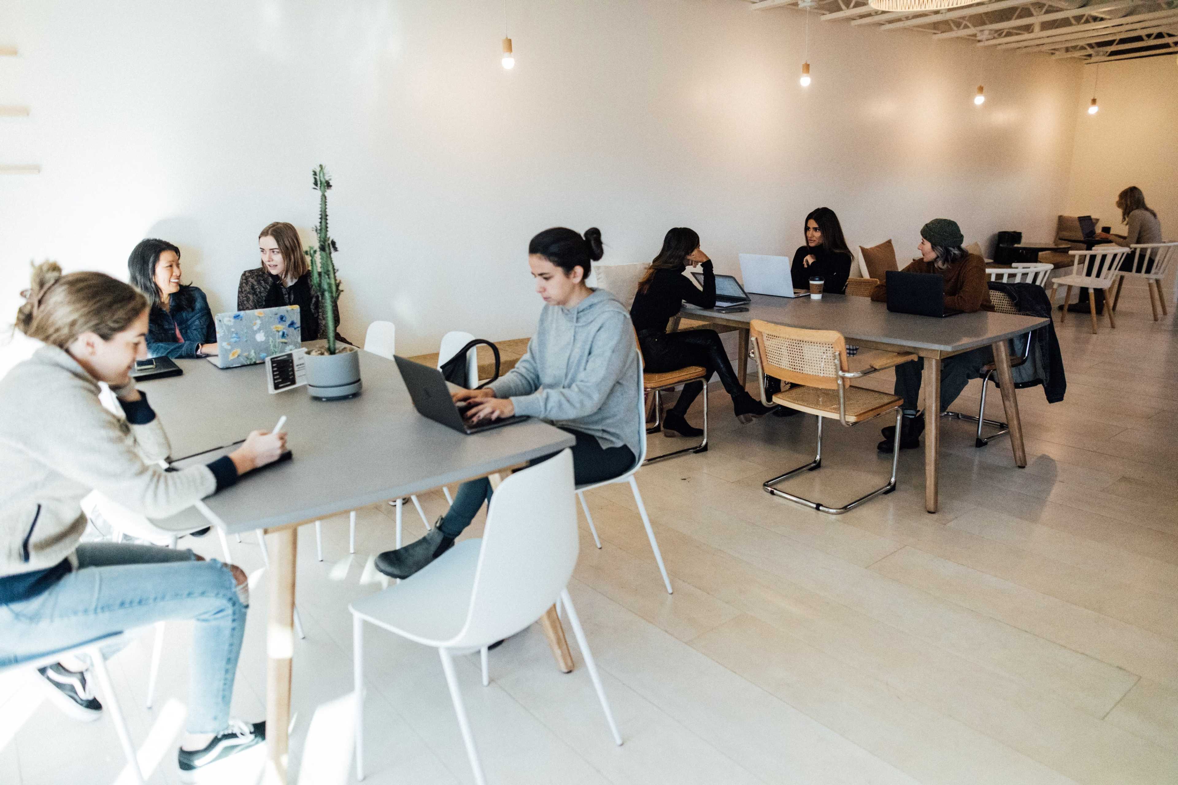 A group of people work on laptops at various tables in a modern, bright workspace.