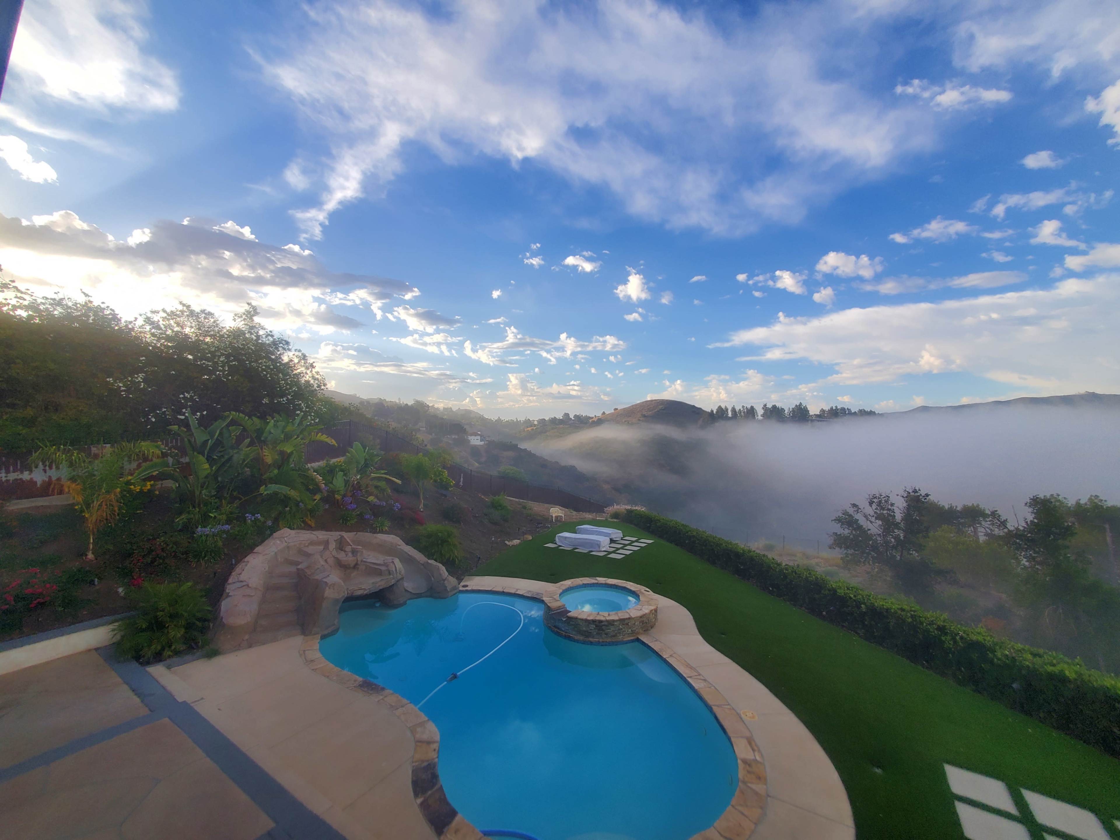 A swimming pool with a stone waterfall is surrounded by greenery and overlooks a misty landscape under a partly cloudy sky.