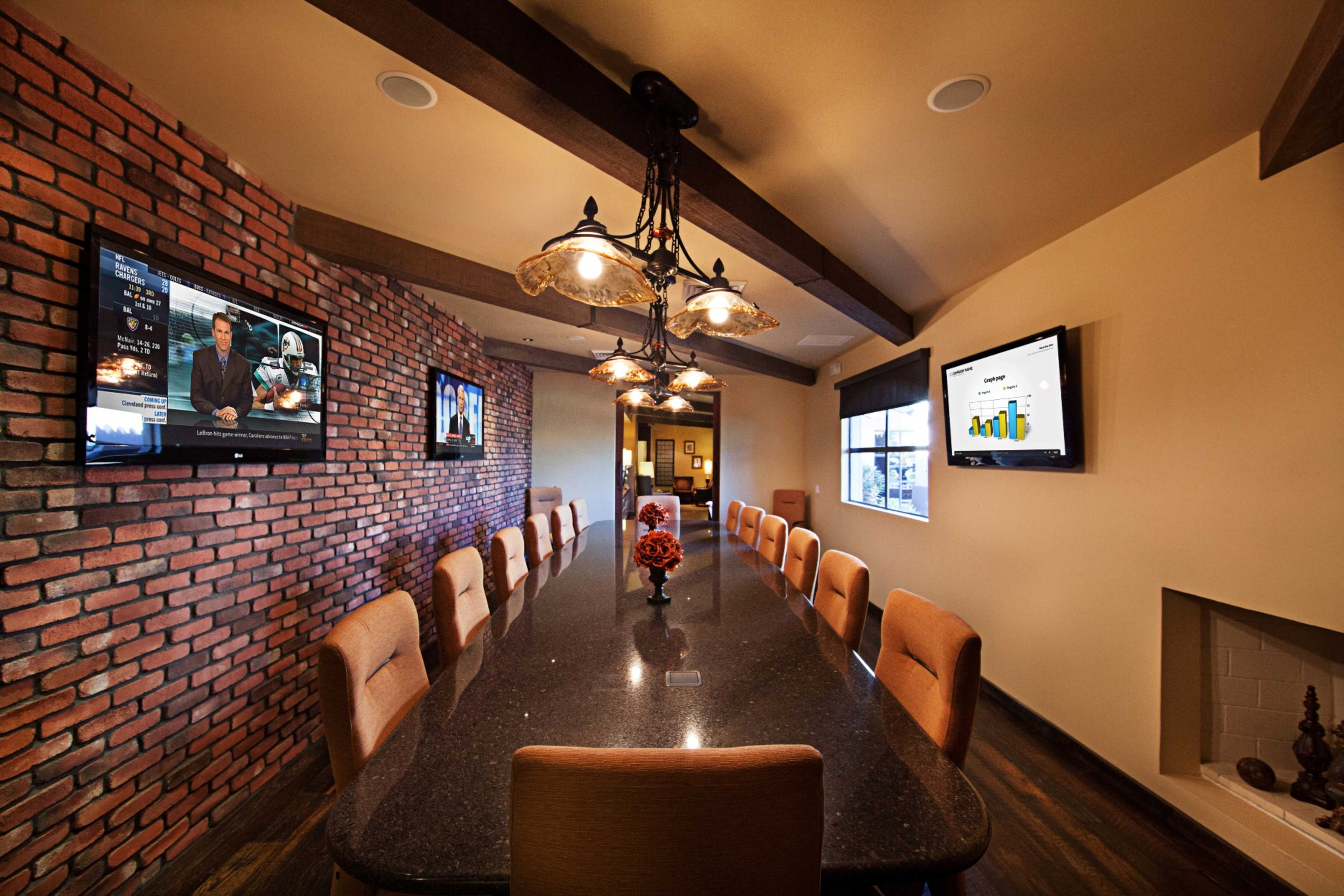 A conference room with a long table, bordered by brick walls and equipped with televisions displaying presentations.