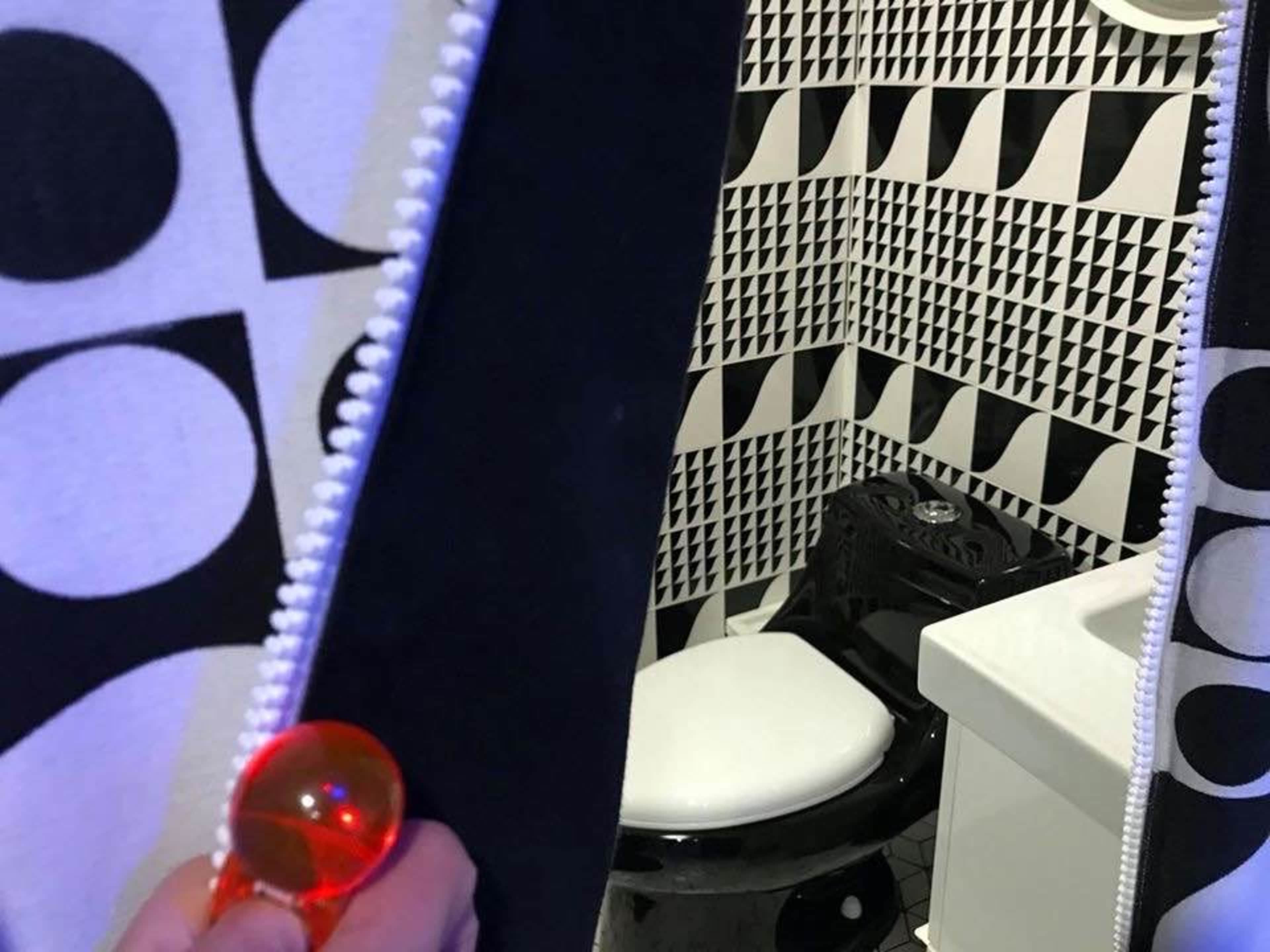 A person holds a red ball while standing in front of a patterned curtain that reveals a bathroom with a black toilet and a white sink.