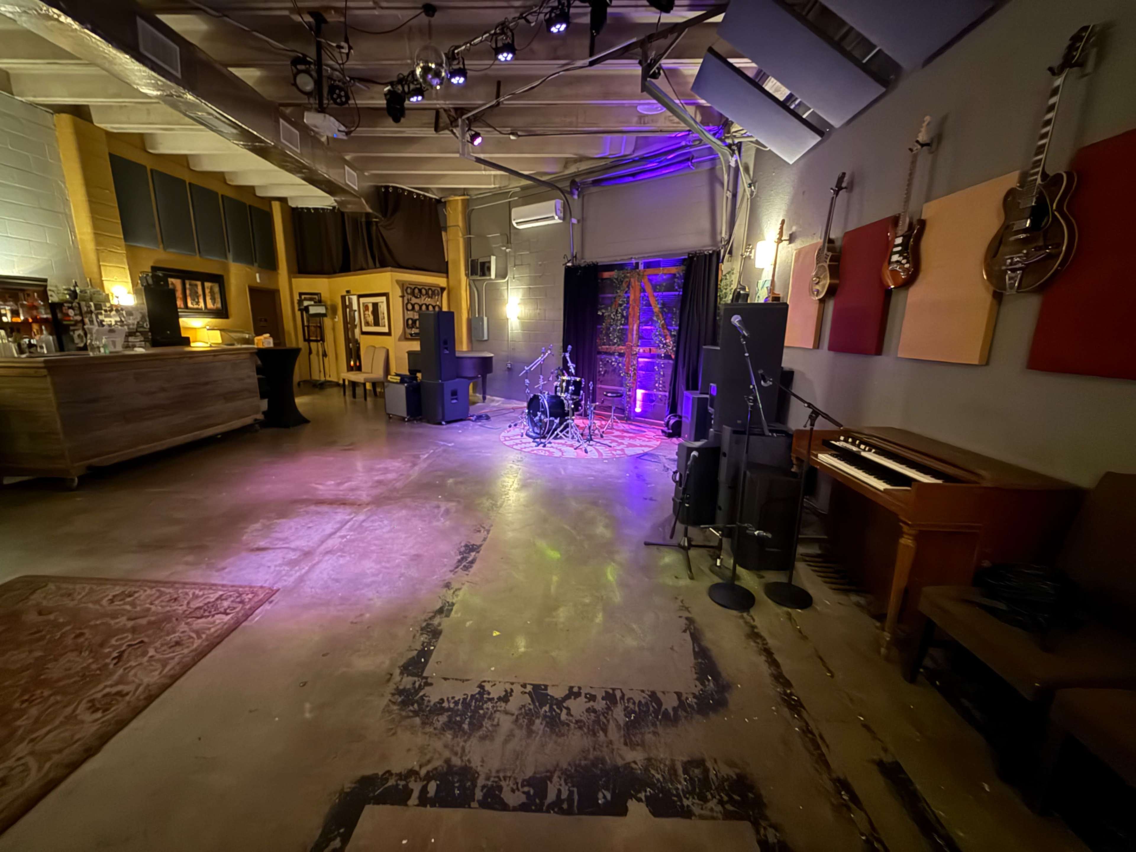The image shows a spacious music venue with a stage set up in the center, instruments, amplifiers, and colorful stage lighting illuminating the area.